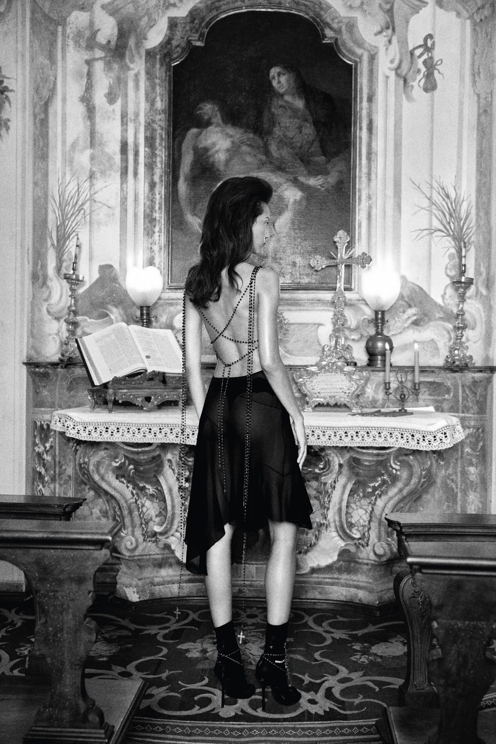 Black rosary-like strands crisscross the bare back and hang to the calves. Black chiffon skirt with an uneven handkerchief hem, sheer through the seat. Black ankle socks. Patent stilettos laced with bead strands, a small cross at one ankle. Pausing at a side chapel altar after everyone else has gone outside.