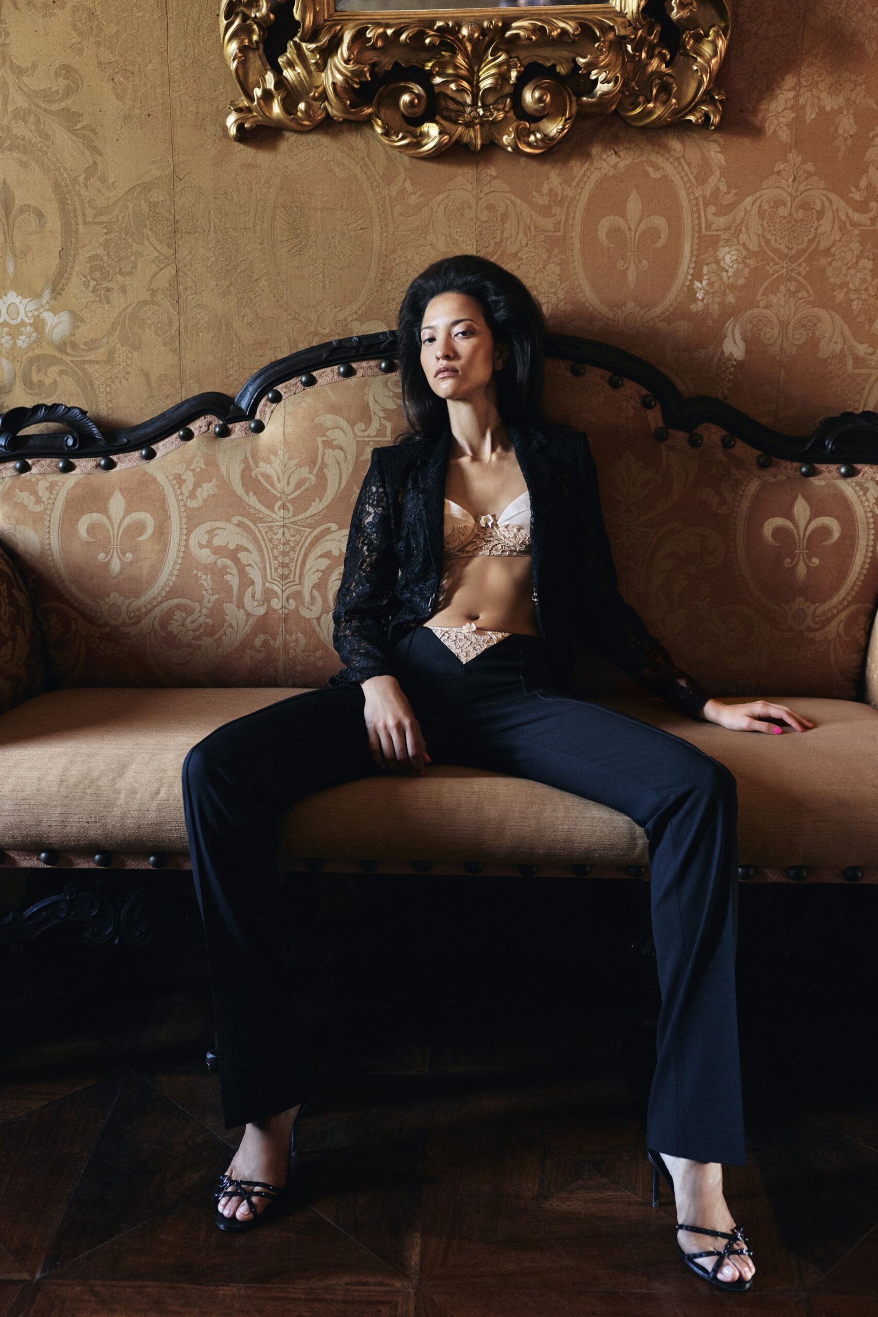 Black lace jacket cut close through the shoulders, left open. Blush satin bra with lace cups and a tiny center bow. Black flared trousers, V-slit waistband filled with matching lace. Black strappy sandals with a small knot over the toes. After the wedding dinner, hiding out beneath a gilt mirror.