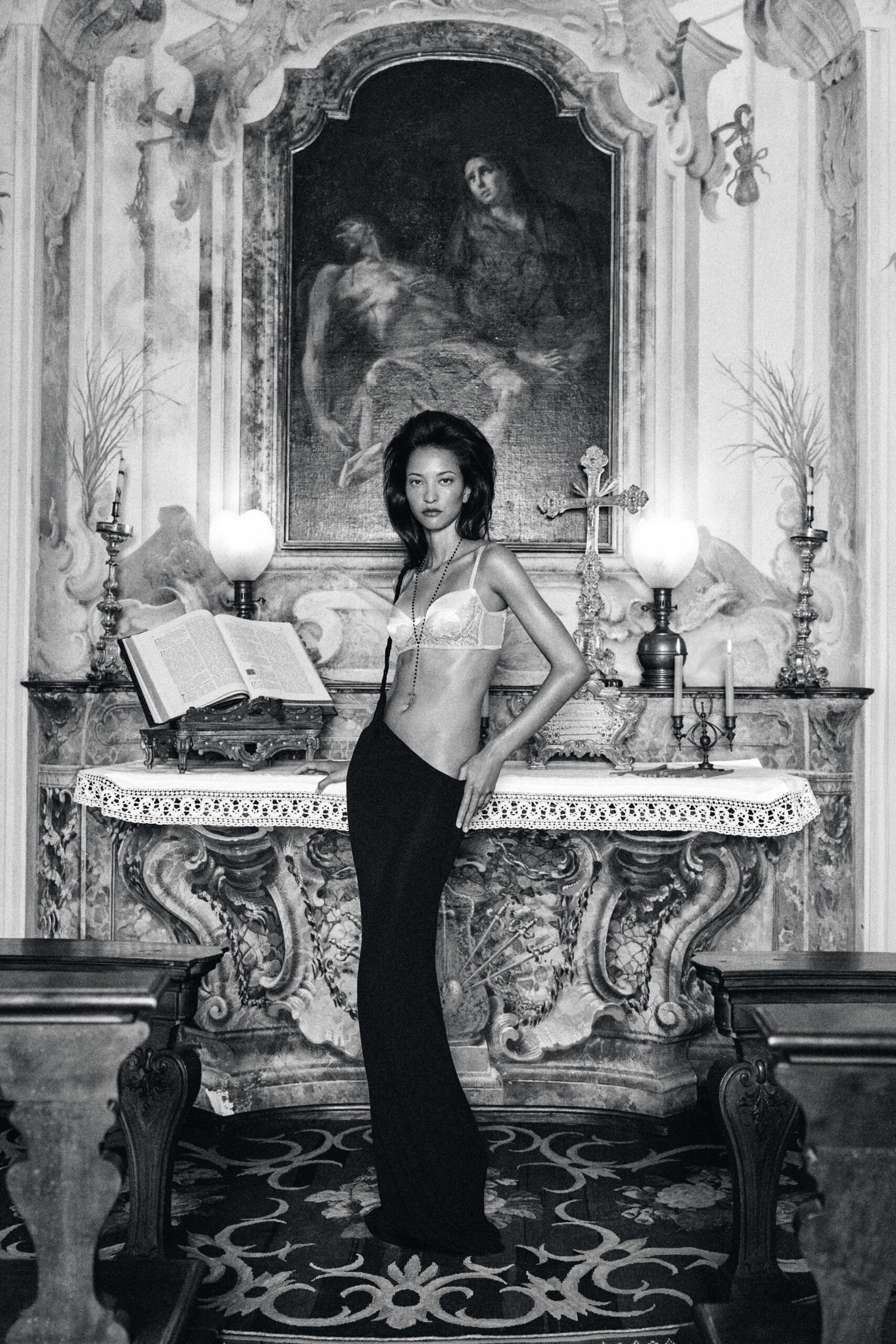 White satin bra with lace cups and narrow straps. Long rosary chain dropping past the navel. Floor-length black skirt cut low across one hip. After the chapel wedding, when the guests have finally gone quiet.