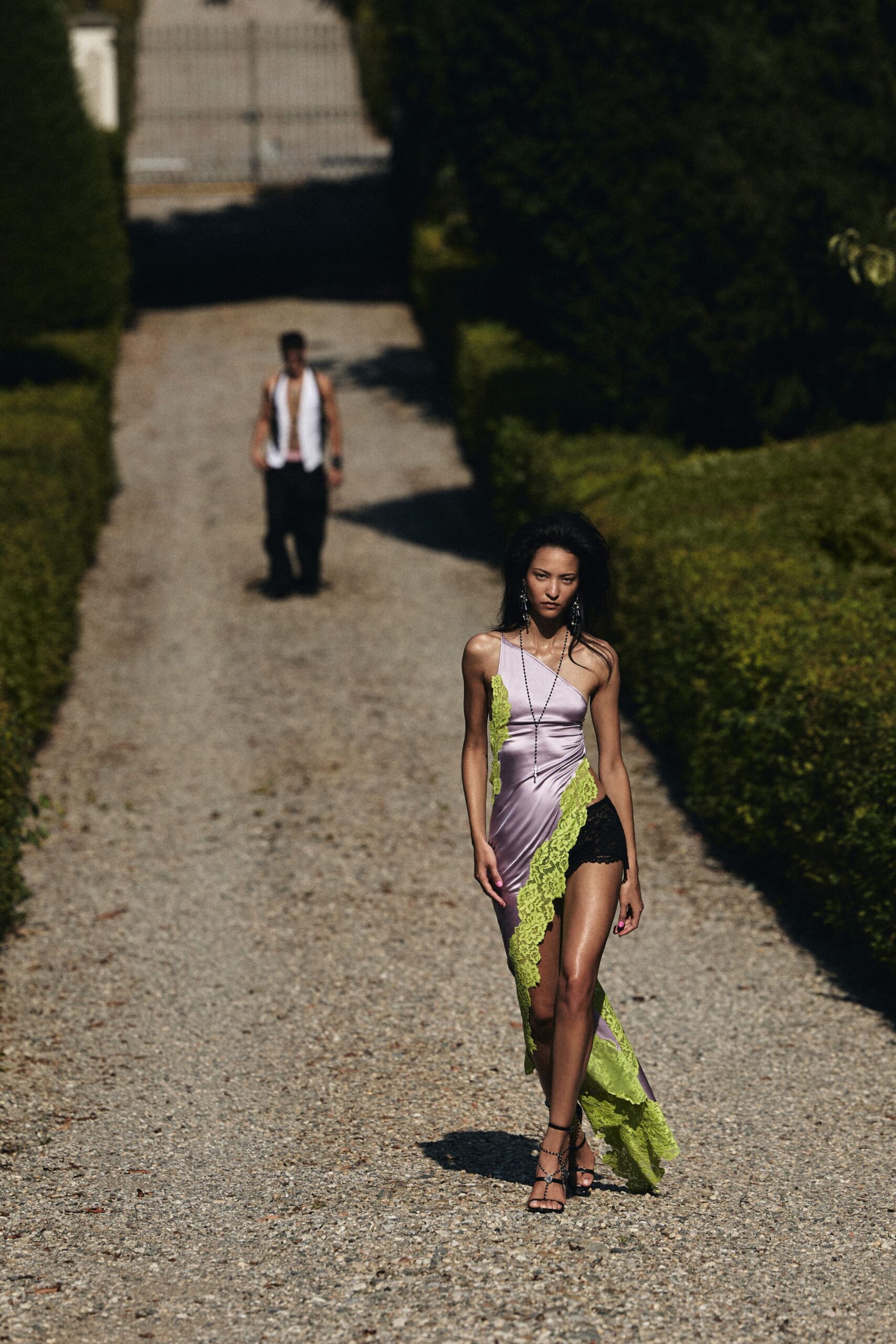 Lilac satin one-shoulder slip dress slashed with chartreuse lace ruffles. Black lace boyshorts exposed through the thigh-high cutaway. Long rosary chain ending in a small cross. Black strappy heels with a toe loop and ankle chain. Crossing a gravel drive after the ceremony went strange in the heat.
