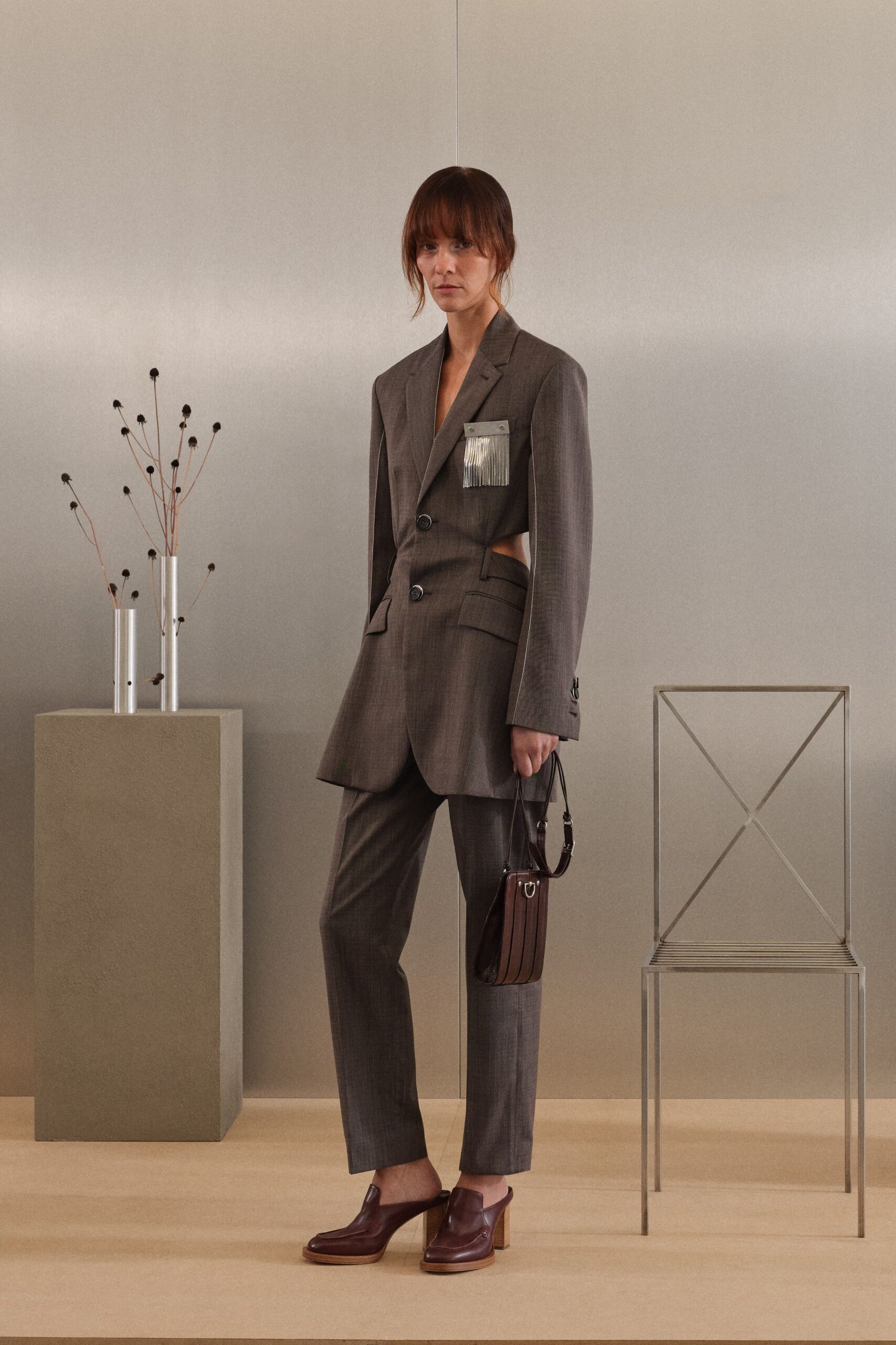 Brown suiting blazer cut away at the waist, with silver fringe pocket trim. Matching straight trousers in the same dry worsted weave. Boxy oxblood leather bag with narrow vertical panels. Oxblood leather mules on stacked wood block heels. For circling a design fair before deciding which chair to buy.