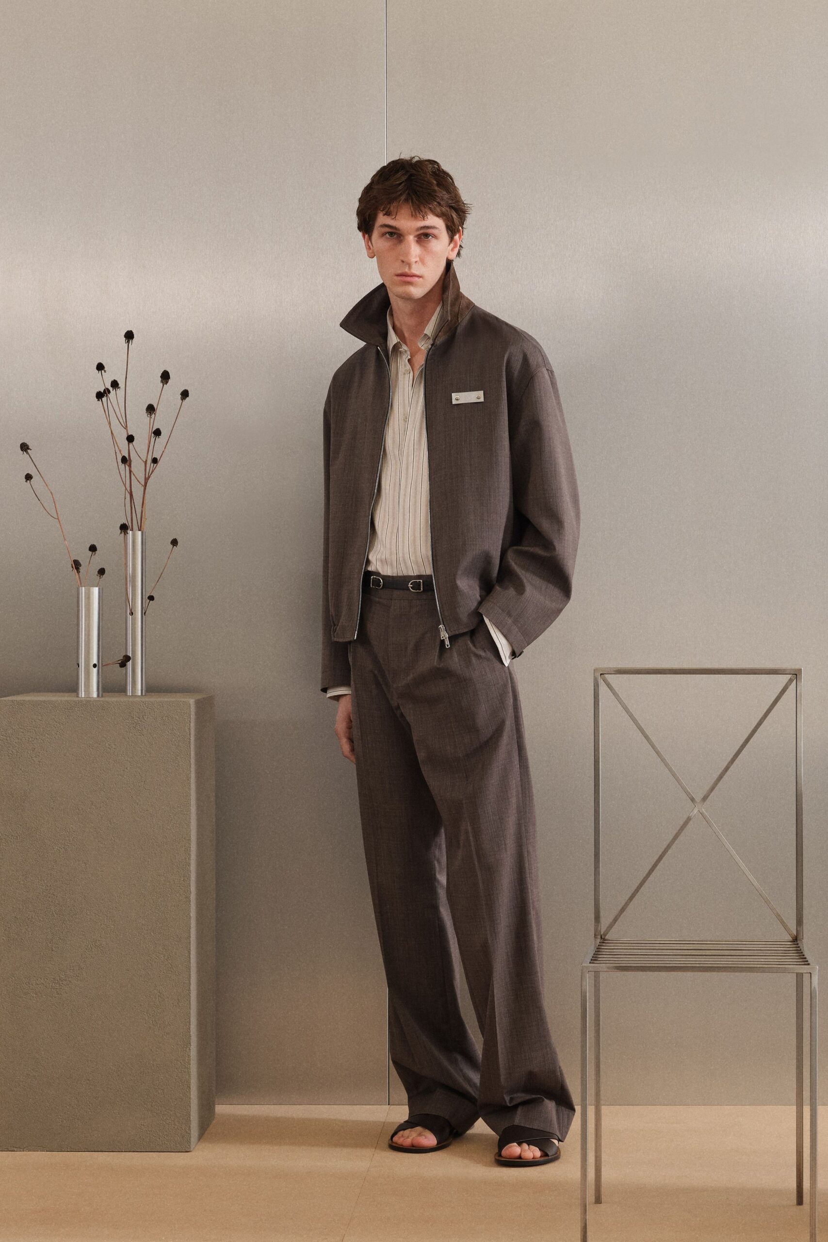 Brown zip jacket with a stand collar and a tiny chest tab. Pale striped shirt, soft placket sitting open at the throat. Matching wide trousers breaking long over the sandal tops. Slim black belt with twin silver loops. Black crossover leather sandals. For drifting through a quiet hotel lobby before breakfast service.