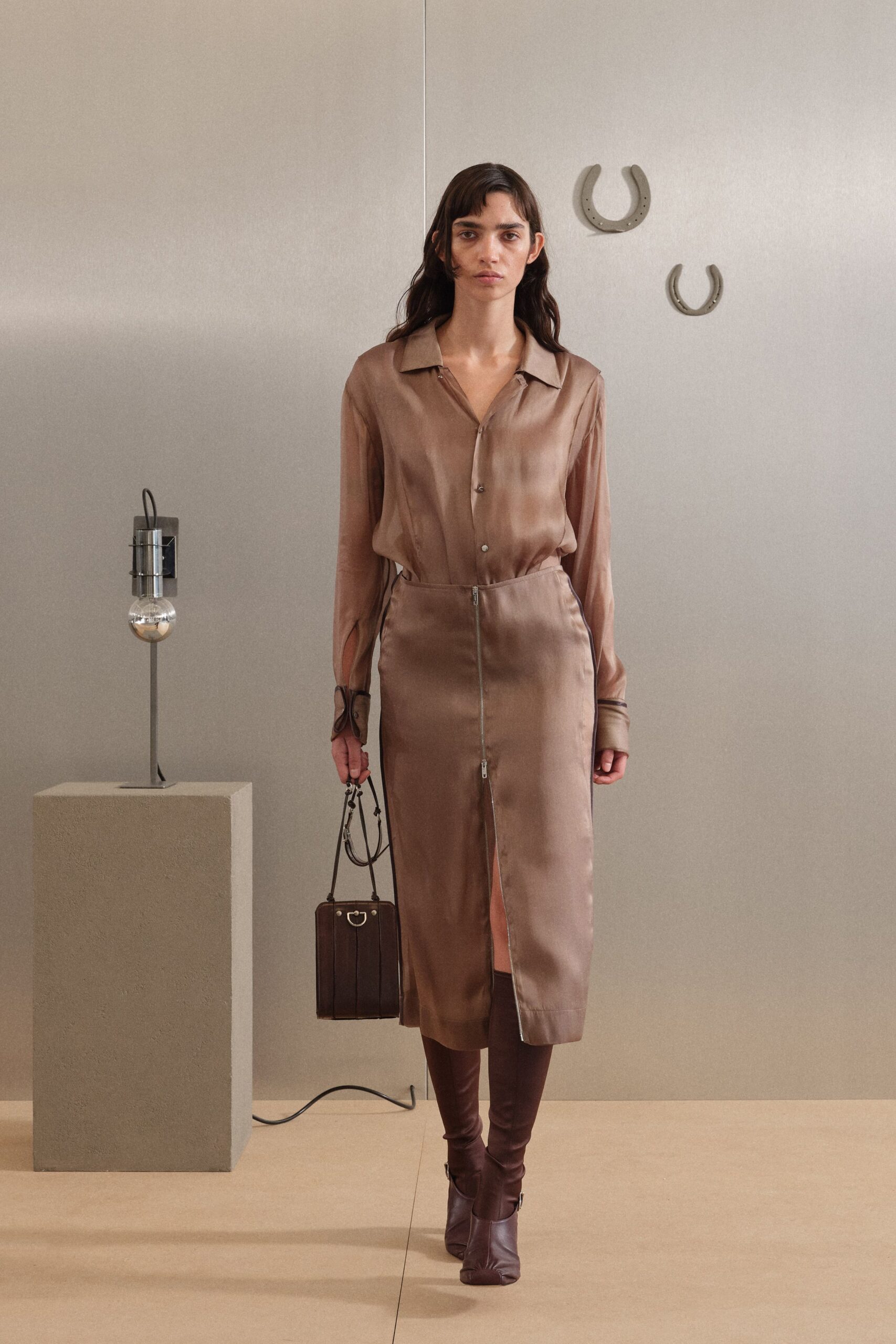 Taupe satin blouse, open collar and long cuffs slit at the wrist. Matching satin midi skirt with a two-way front zip opening the hem. Boxy espresso leather bag with a small D-ring tab. Dark chocolate leather boots, softly ruched through the shin. For checking into a quiet hotel before the wedding guests arrive.