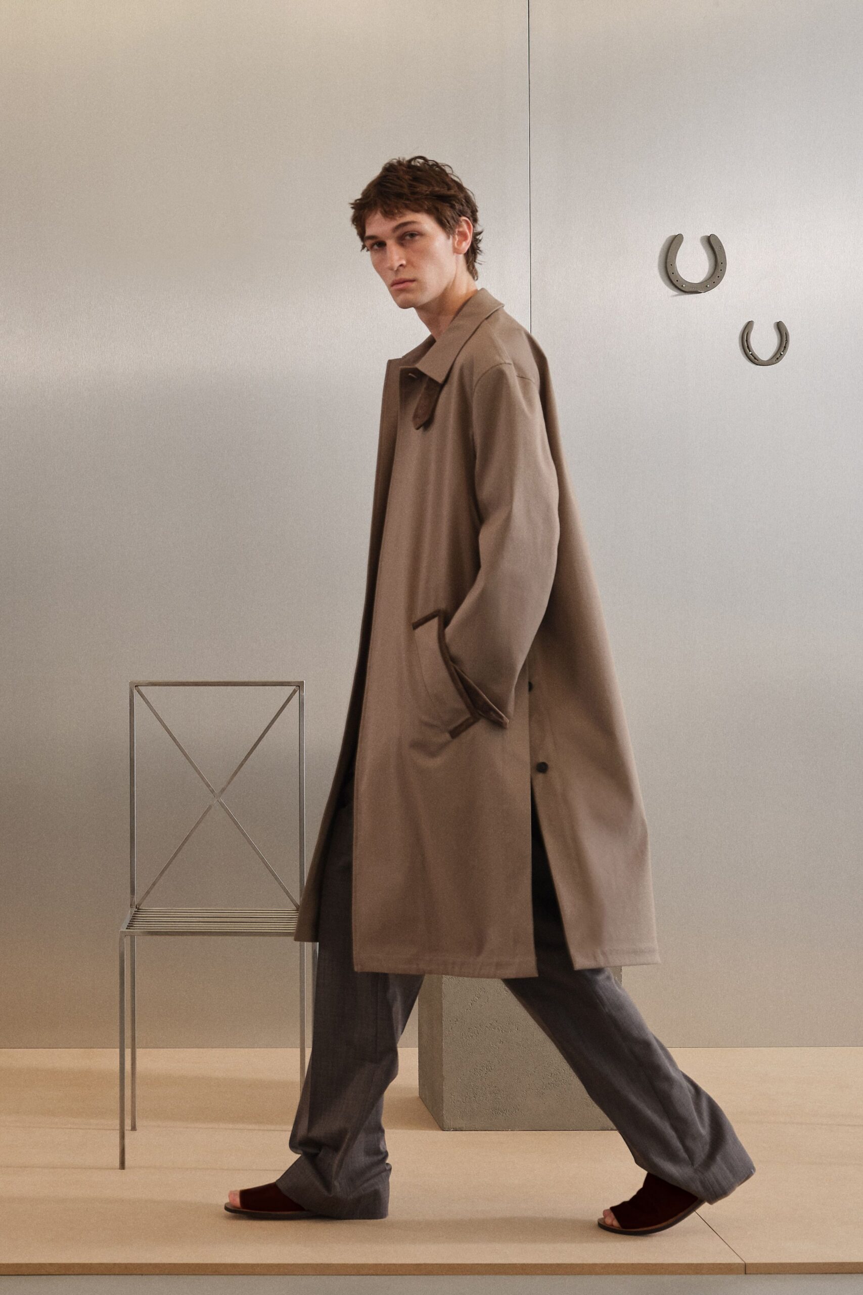 Taupe single-breasted overcoat with dark leather trim at collar and pocket. Slate trousers falling loose with a soft break over the foot. Oxblood velvet open-toe mules, flat and barely clearing the floor. Crossing a quiet hotel lobby before breakfast service starts.