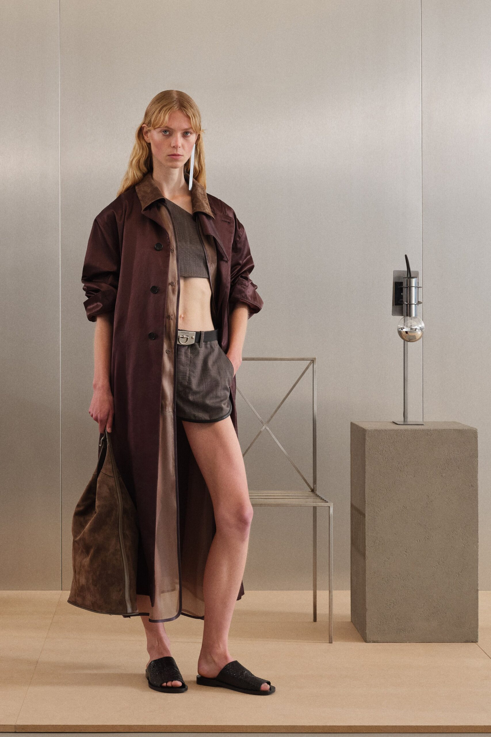 Burgundy satin trench, taupe sheer placket hanging loose beneath. Brown gauze crop top cut blunt above the ribs. Grey micro shorts with black binding and a square silver buckle. Slouching tobacco suede tote with a long exposed zip. Black woven leather slides. For the first drink in a hotel bar before anyone else arrives.