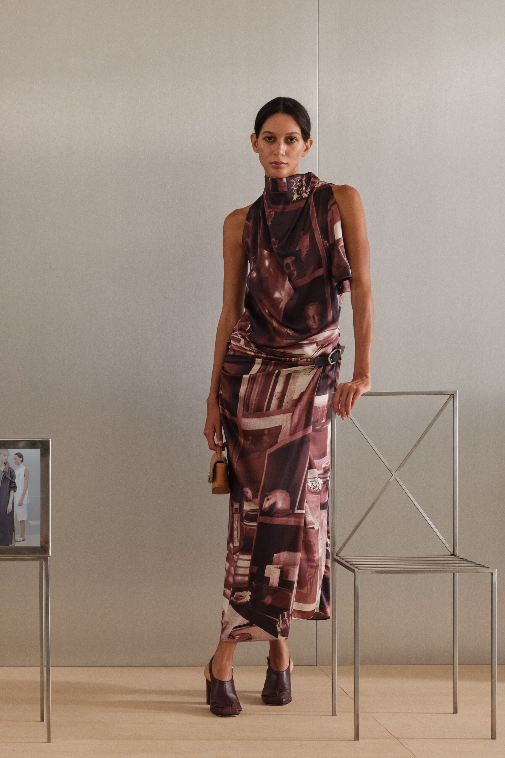 Burgundy satin column dress, photo-print panels draped into a scarf neck. Brown leather hip belt with an oval silver buckle. Tiny tan box bag. Eggplant leather mules, squared and softly creased at the vamp. At a hotel lounge before dinner, still carrying the room key.