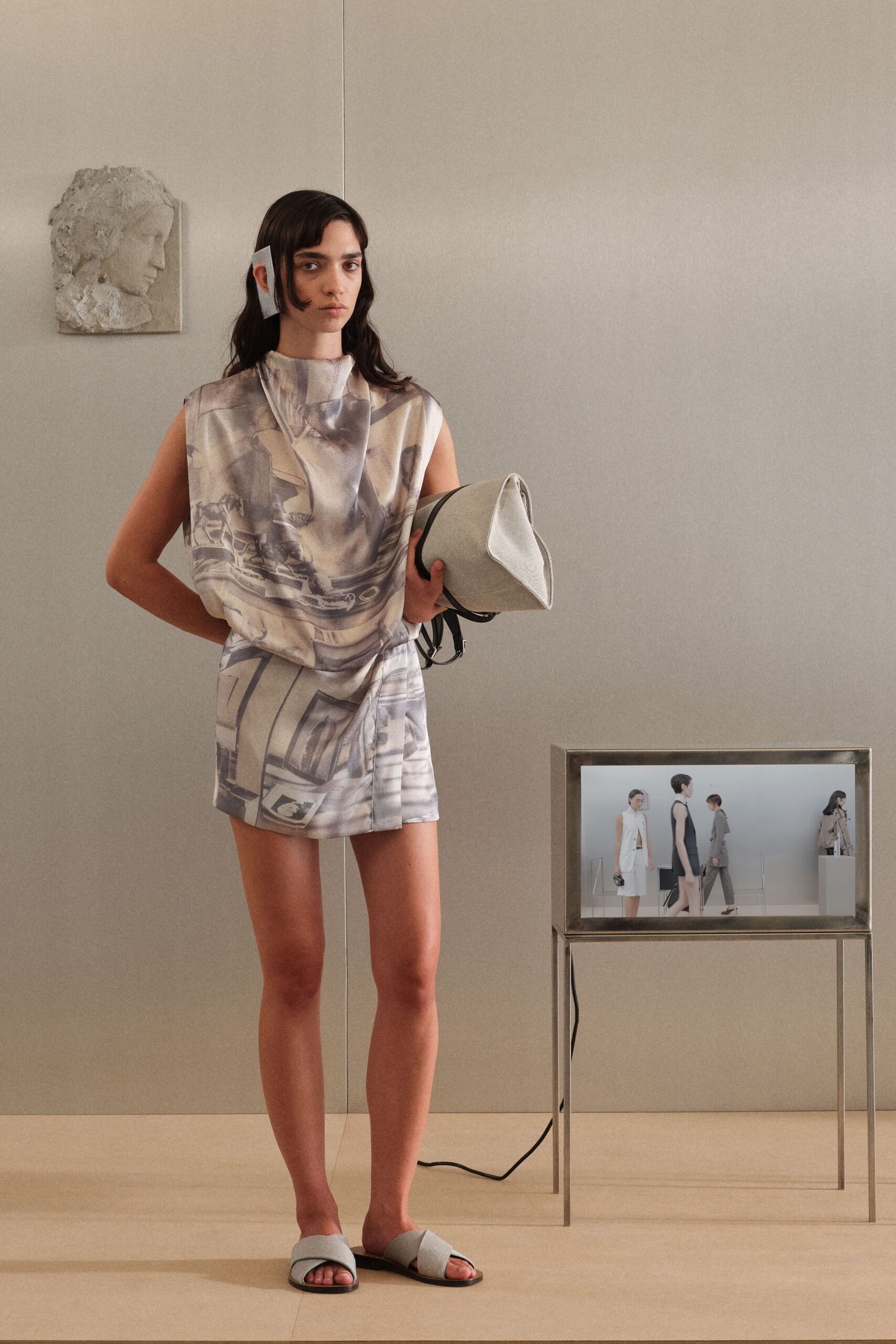 Translucent rectangular clip pinned flat against one ear. Sleeveless dove-grey satin mini, softly draped with a blurred photographic print. Pale canvas bucket bag with a white folded leather corner. Ash fabric crisscross slides on black soles. At a quiet gallery install, carrying the catalogue before the doors open.