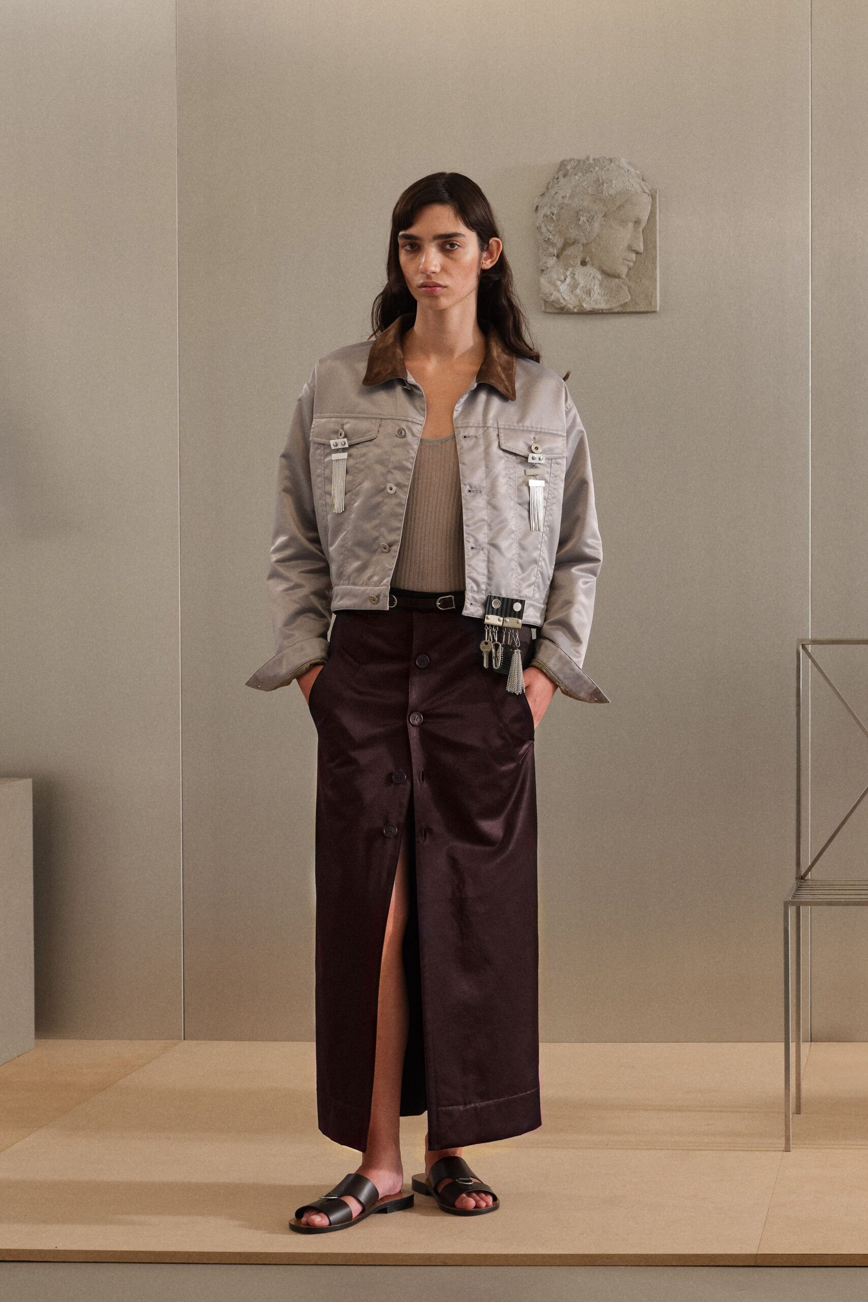 Pearl-grey satin jacket with a brown suede collar and dangling metal tassels. Taupe ribbed tank, cut narrow through the chest. Deep aubergine satin column skirt, buttoned down the front with a long slit. Black leather slide sandals with a plain toe strap. Museum hush, killing ten minutes before the courtyard lunch.