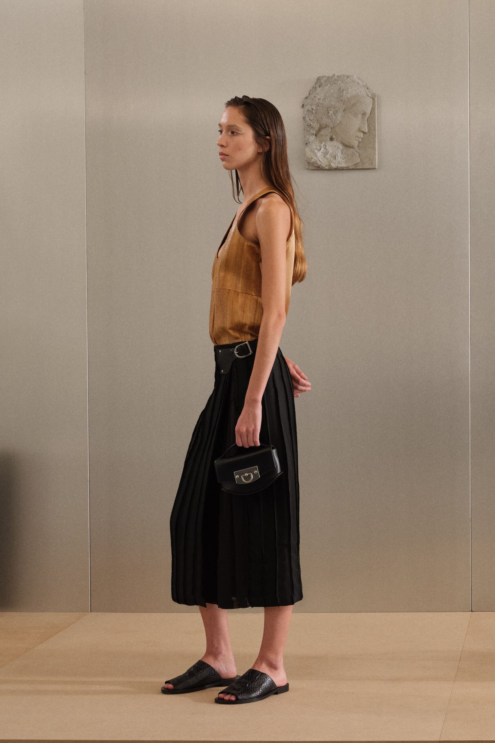 Ochre suede sleeveless top with a deep V and panel seam. Black pleated midi skirt, cinched with a side buckle strap. Small black top-handle bag with a horseshoe clasp flap. Black woven leather slides. Museum hush, half an hour before the donors arrive.