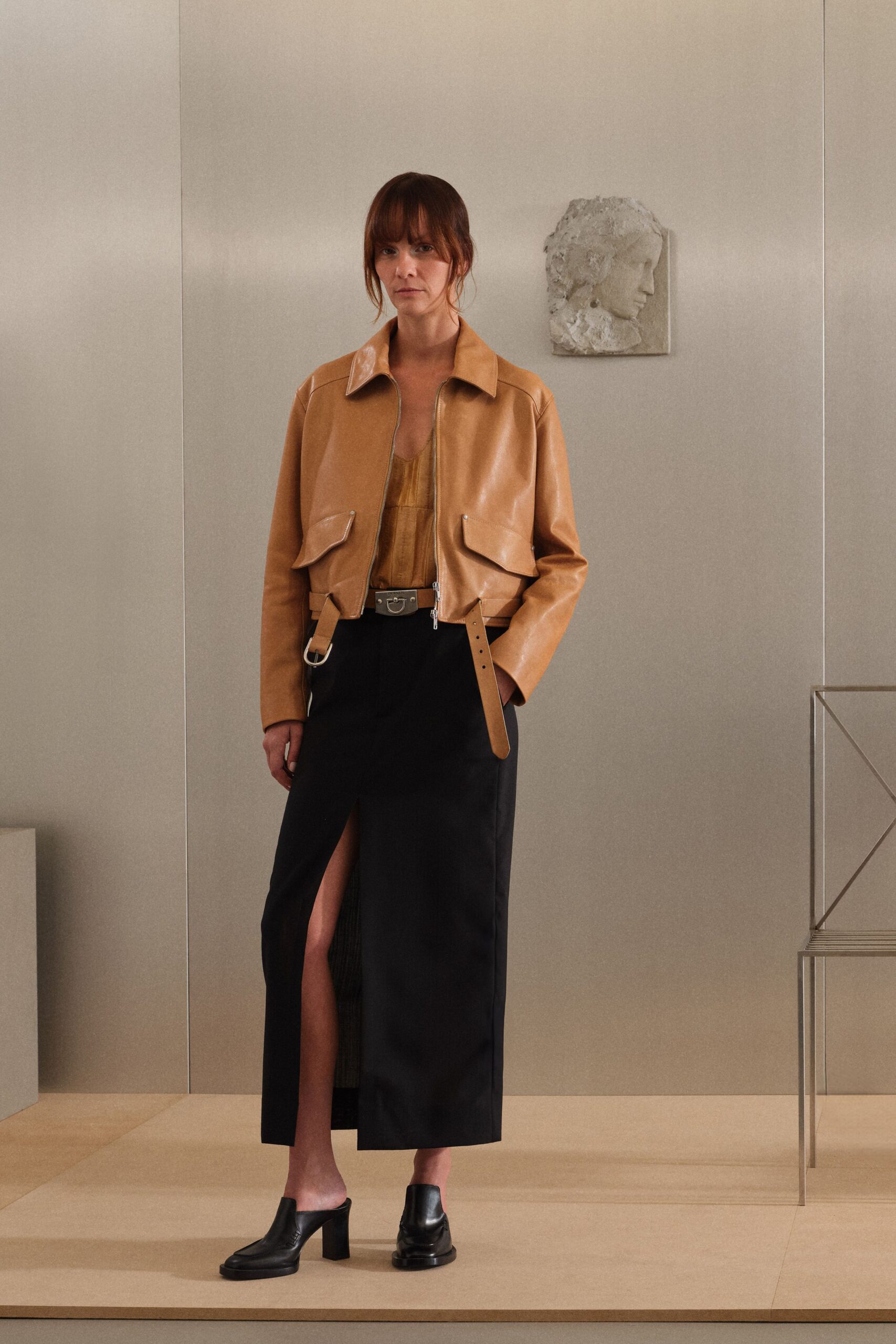 Cognac leather jacket, cropped blunt with hanging buckle tabs and D-rings. Burnished ochre camisole with a raw, softly crumpled sheen. Black column skirt split high at the front, one panel pleated beneath. Black leather mule clogs on a square stacked heel. For crossing a quiet sculpture court after the opening crowd has gone.