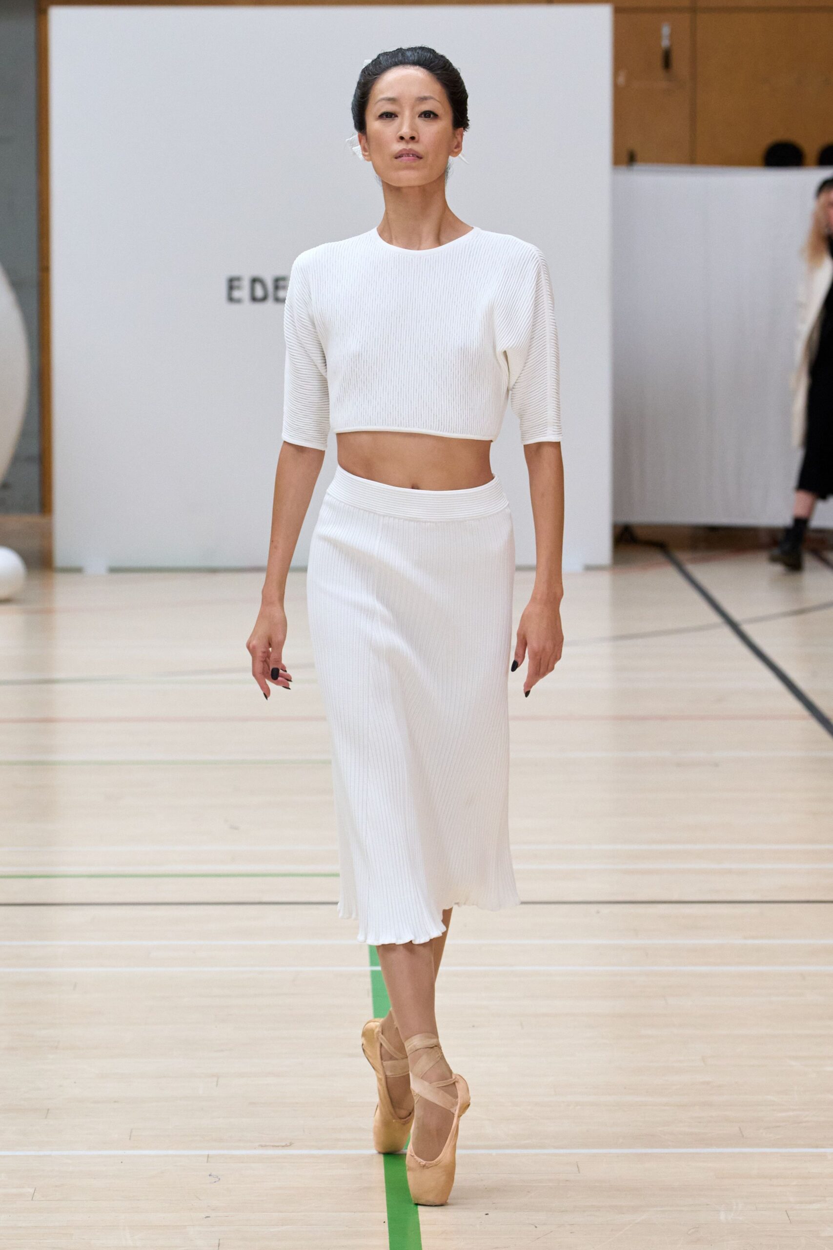 White ribbed knit crop top with elbow sleeves and a clean rolled hem. Matching ribbed knit midi skirt, waistband sitting high above a softly fluted edge. Blush satin pointe shoes crisscrossed with ribbons. Crossing a gym floor after the recital, still half inside the costume.