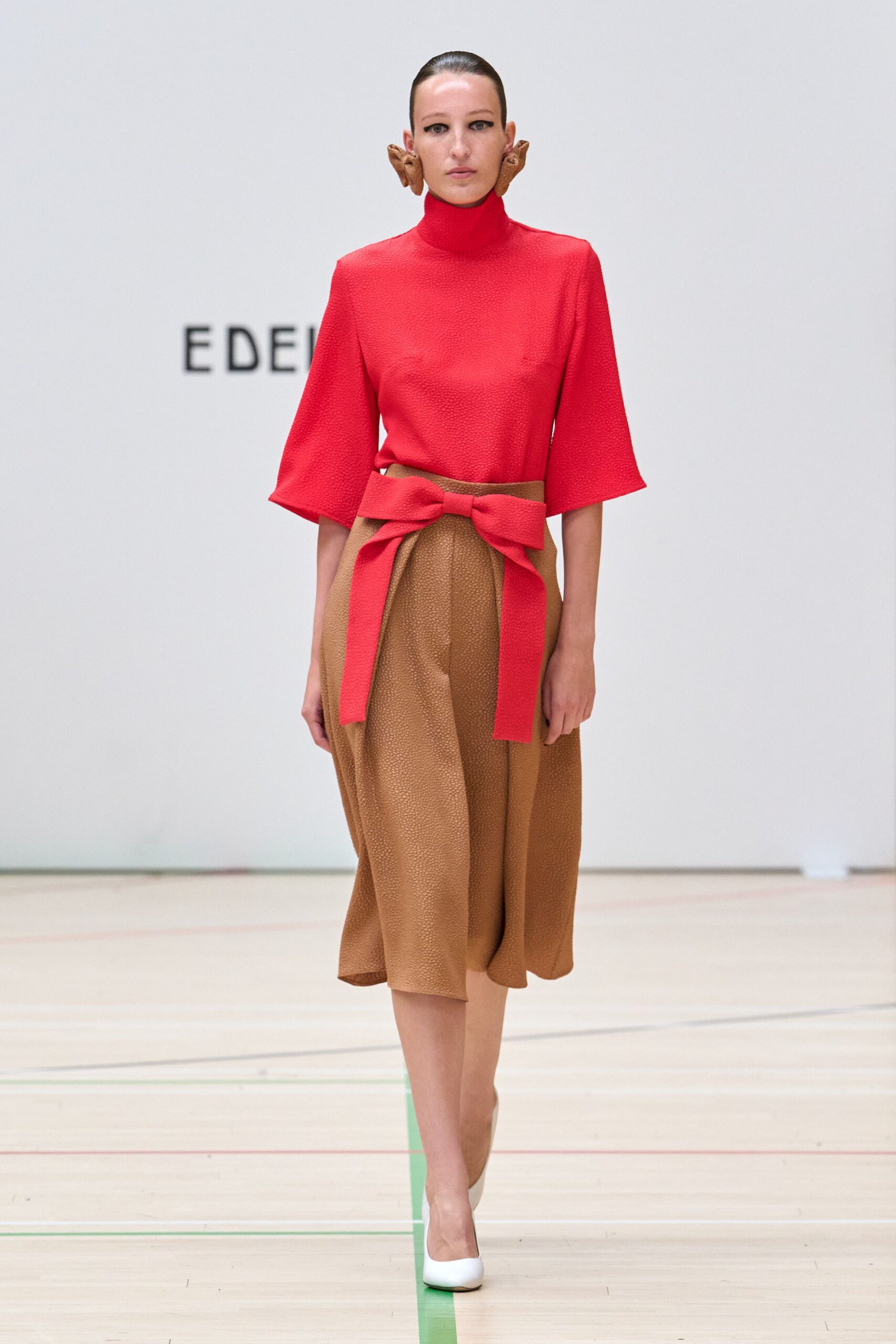 Tomato red pebble-textured top with a high rolled neck and wide sleeves. Camel pebble-textured midi skirt, front split hidden under a broad tied sash. Carved wooden spiral earrings hugging the jaw. White pumps with blunt almond toes. Museum atrium at noon, still holding the program from a craft fair.