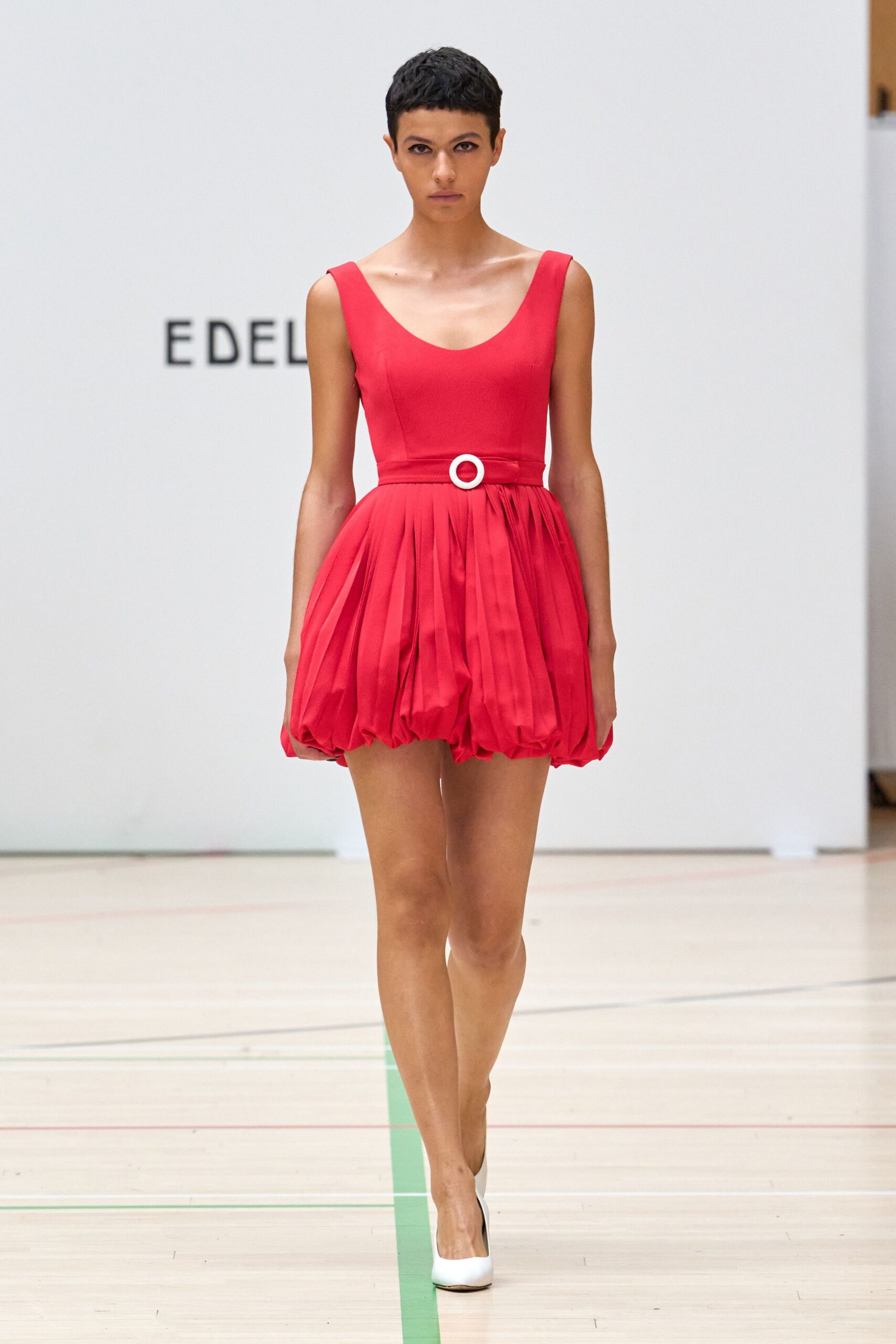 Tomato red mini dress with a scooped neck and ballooned pleated hem. Self belt cinched with a white circular buckle at the waist. White leather pumps, sharply pointed from the front. Walking across a school gym floor after the chairs have been cleared.