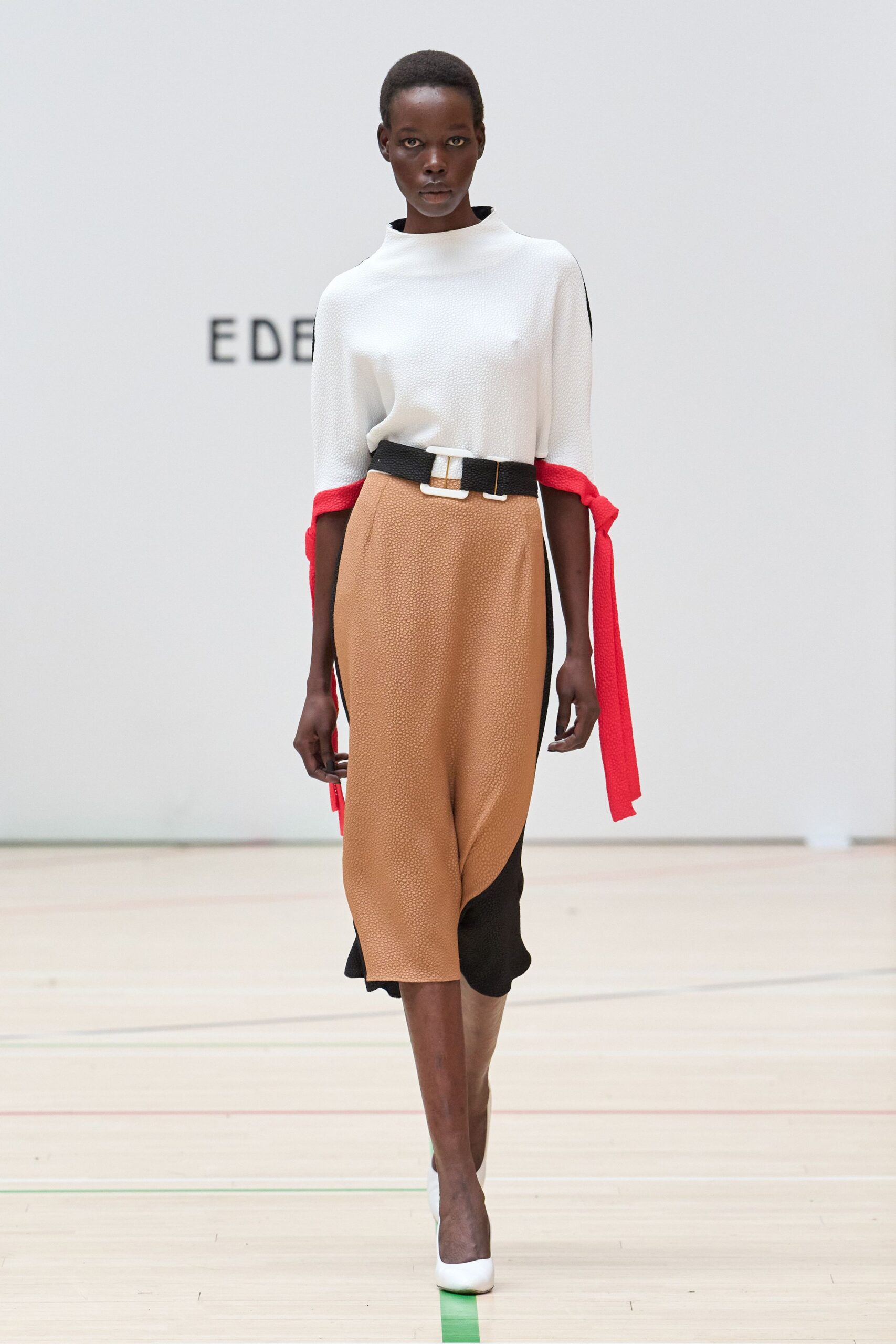 White pebble-textured top with a funnel neck and red tied cuffs. Camel pebbled midi skirt, black side panels curving under the hem. Black woven belt with a square white buckle. White pumps. Crossing a museum atrium before the school groups arrive.