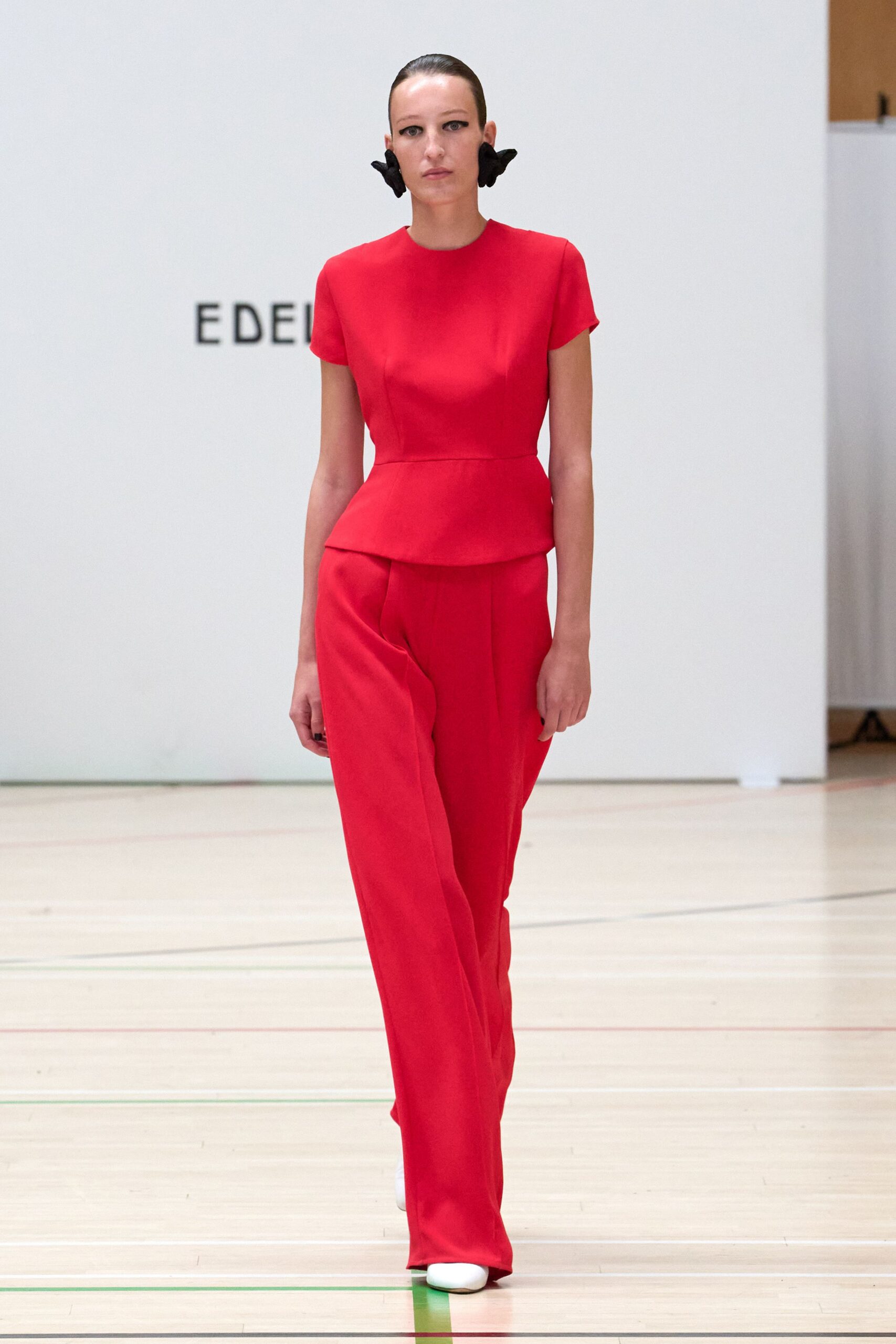 Scarlet crepe top with cap sleeves and a curved peplum flare. Matching straight trousers with a pressed center crease breaking at the shoe. Black sculpted flower earrings hugging both earlobes. White rounded-toe boots. Crossing a varnished gym floor for the prizegiving nobody dressed down for.