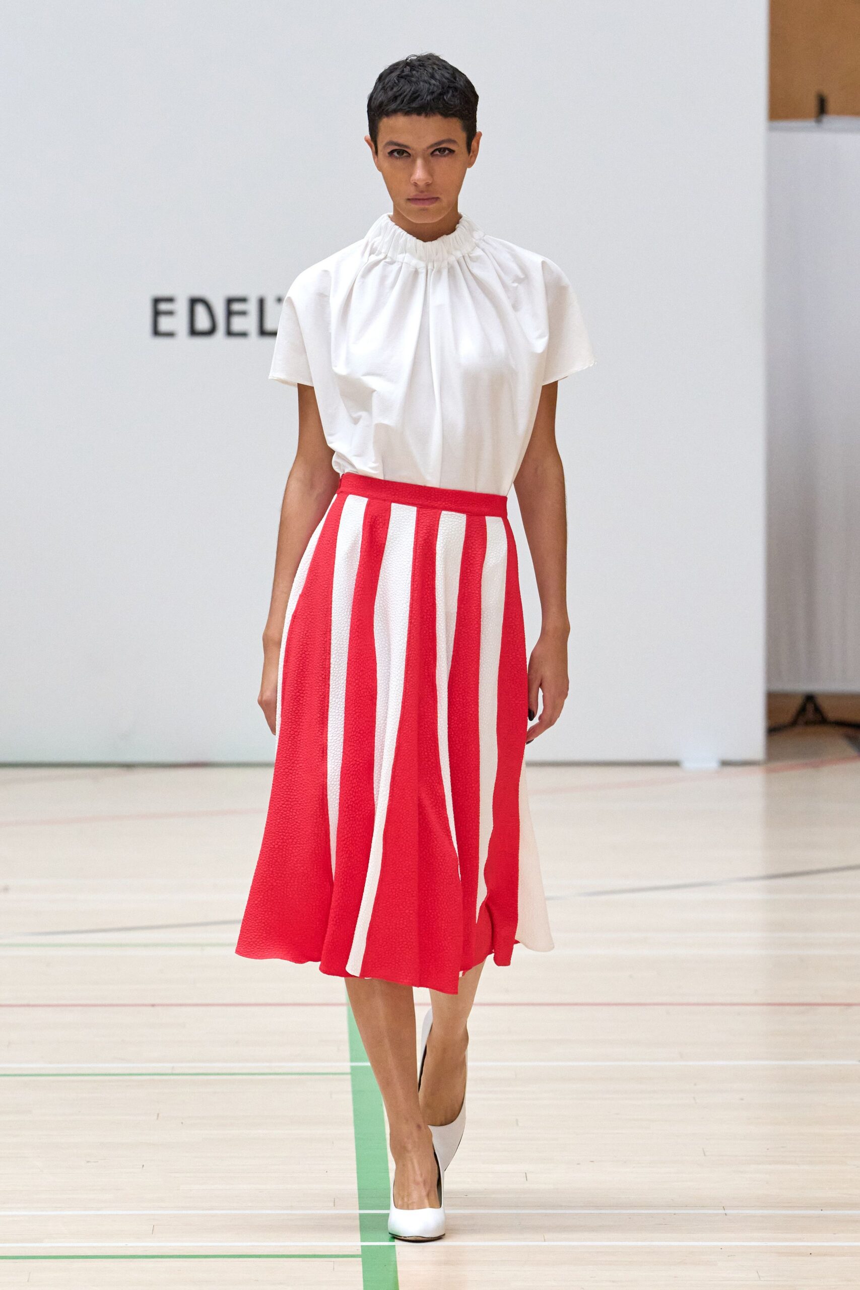 White blouse in crisp poplin, neckline gathered into a ruffled funnel. Red and white striped midi skirt in pebbled knit, hanging in soft flares. White pumps with a blunt vamp. Walking into a civic atrium just before the speech starts.
