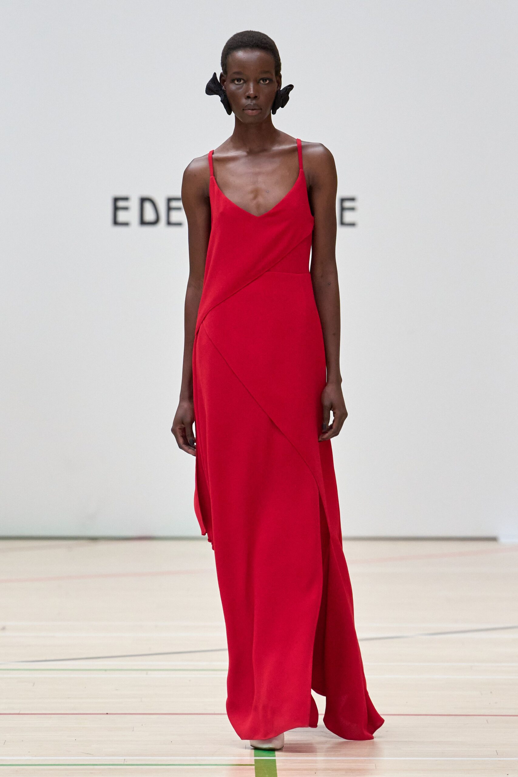 Scarlet slip gown in matte crepe, cut with a sweeping diagonal overlay slit. Black bowlike earpieces hugging both sides of the head. Crossing a school gym floor after everyone else has gone home.