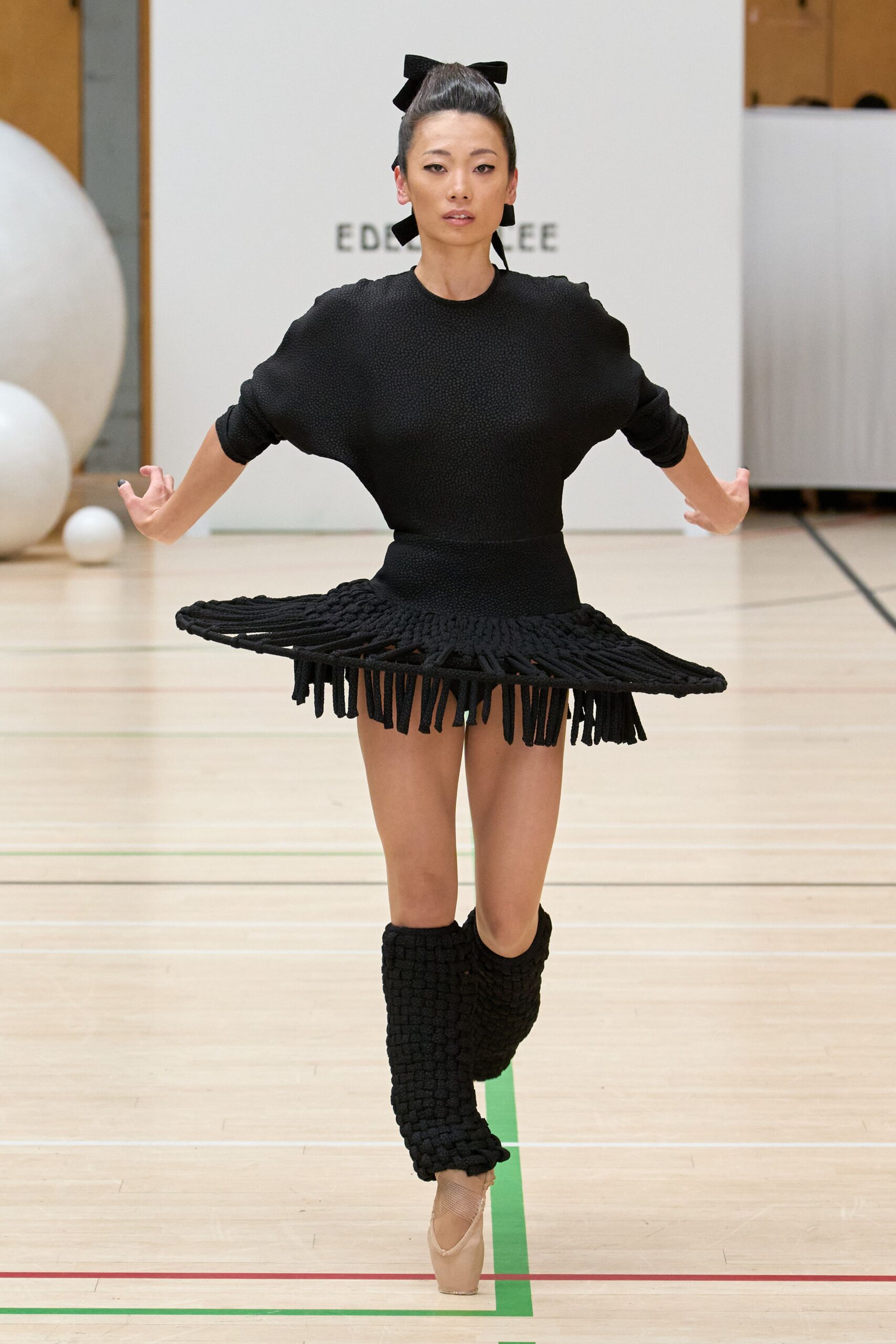 Black grosgrain bow pinned above a lacquered updo. Black pebble-textured knit dress with a stiff rope-fringed pannier skirt. Chunky basket-woven black knit rising almost to the knee. Blush satin pointe shoes with crisscross ribbons at the instep. Center court before the recital, gym floor lines still underfoot.