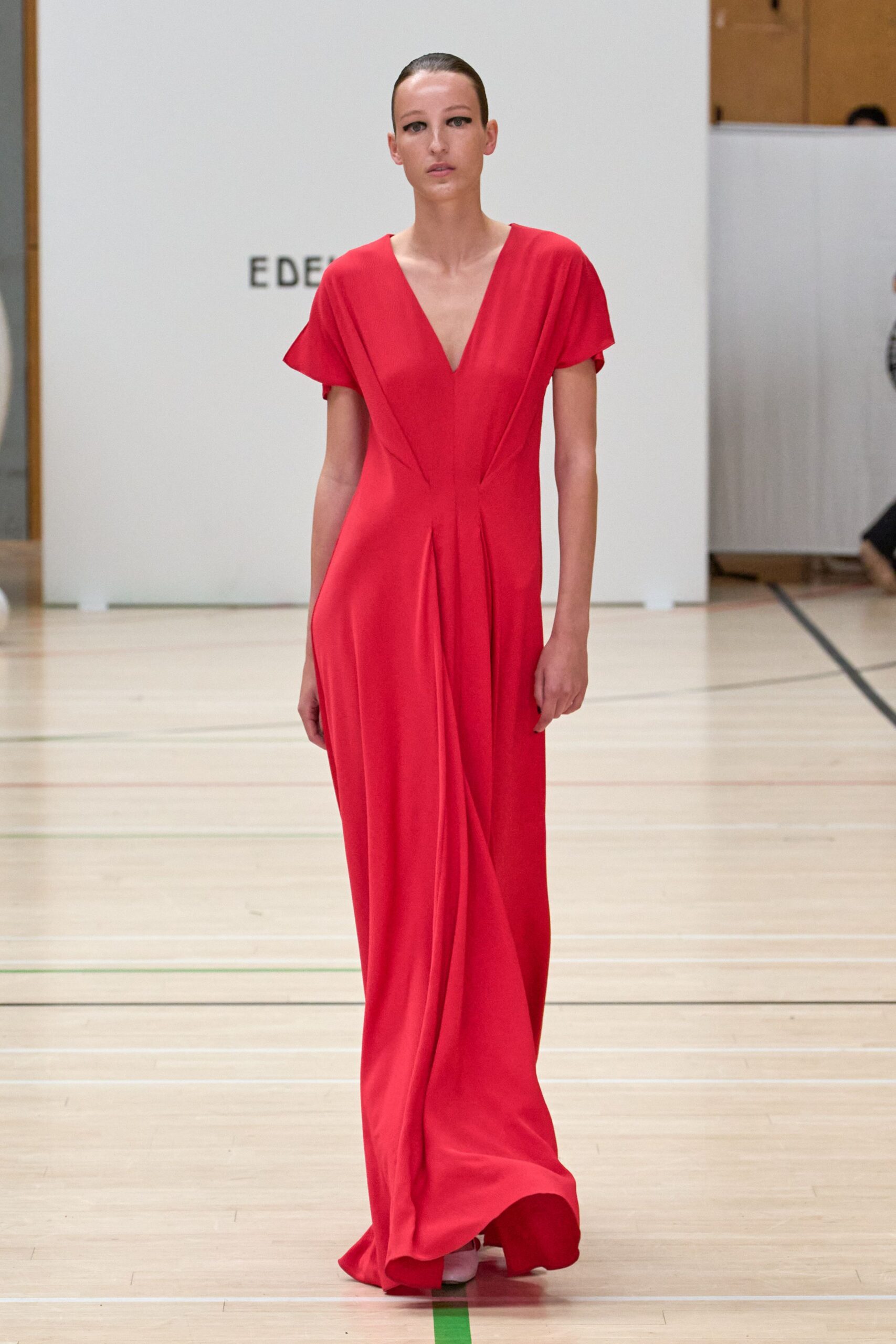 Tomato-red crepe gown with a deep V and soft waist pleats. White pointed heels barely visible under the dragging hem. Crossing a gym floor after midnight, still dressed for a harder room.