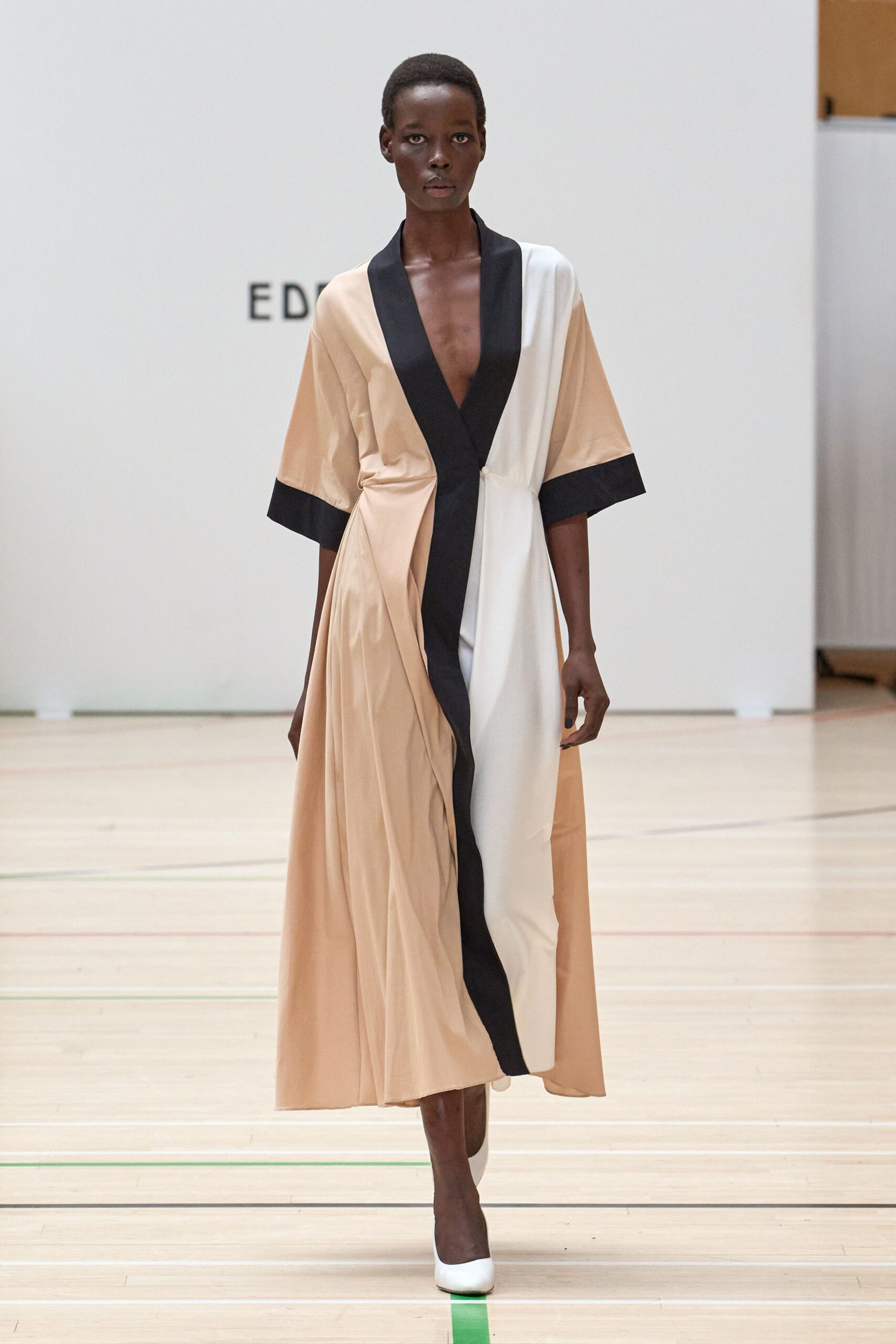 Fluid satin wrap dress in biscuit and ivory, black shawl placket cutting to the waist. White pumps with a blunt vamp. Crossing a gym floor after the folding chairs have been cleared.