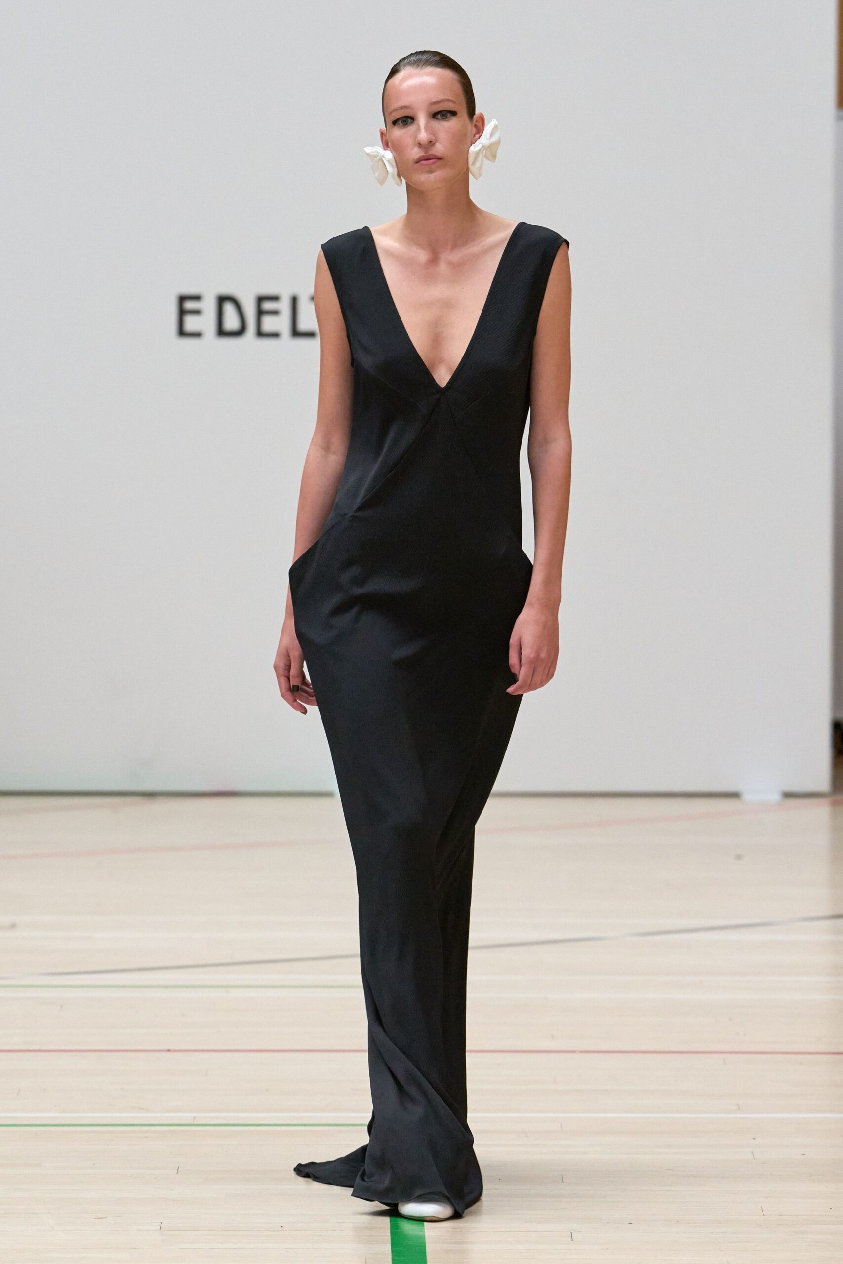 Oversized white fabric bows knotted close against each ear. Black floor-length gown in matte satin, deep V and slanted seam-tucked pockets. White rounded shoes peeking from the pooled hem. Walking across a waxed gym floor after everyone else has gone home.