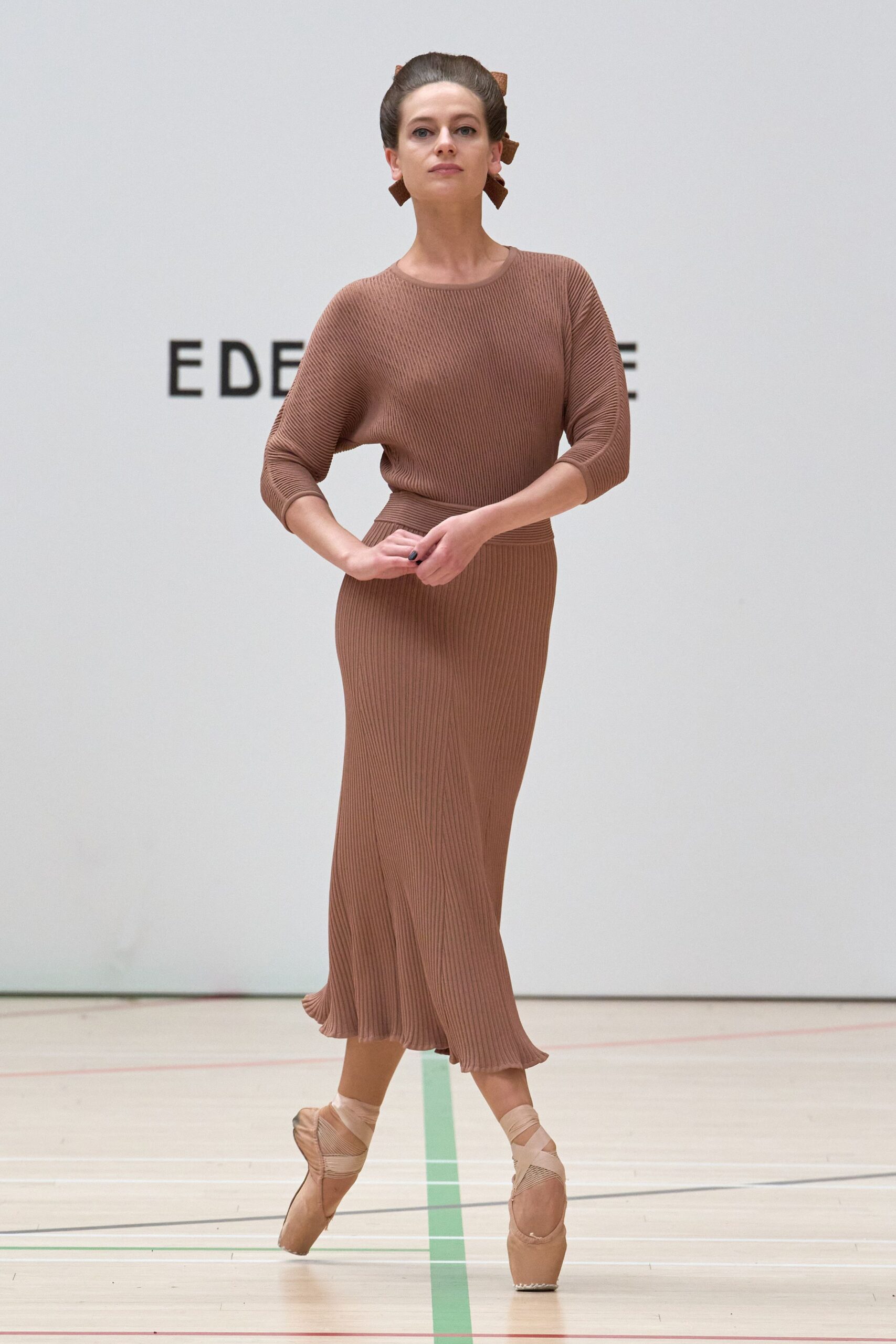 Cocoa ribbed knit top with batwing sleeves and a close crew neck. Matching ribbed midi skirt, hem loosening into a soft flutter. Blush satin pointe shoes with crisscross ribbons at the ankle. School gym floor, half an hour before the recital parents arrive.
