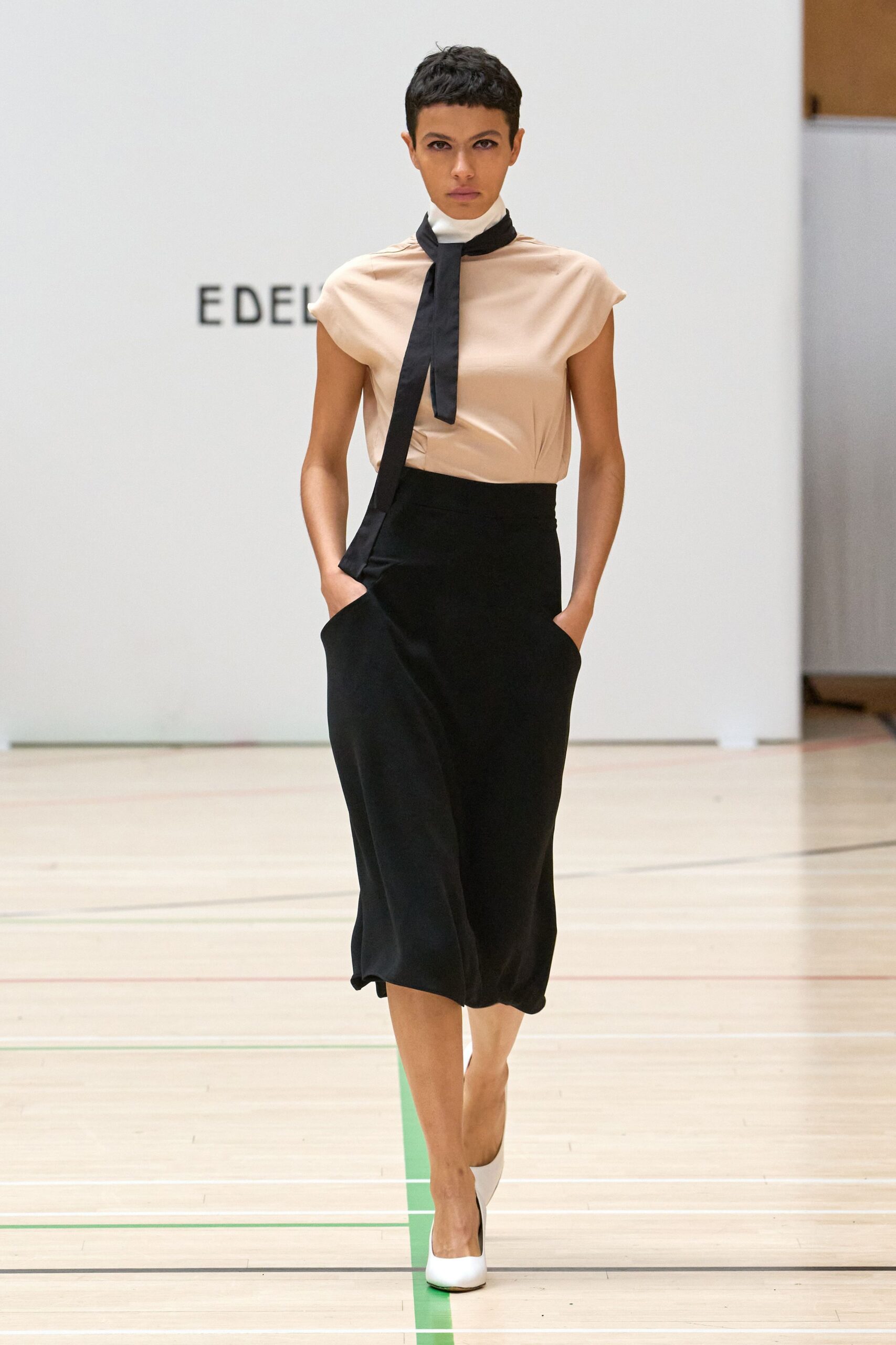 Blush satin cap-sleeve top, softly bloused under a white funnel neck. Black neck scarf hanging in two long matte tails. Black column skirt with deep hip pockets and a slightly pooled hem. White pumps with squared toes. Crossing a gym floor after the speeches, still dressed for somewhere else.