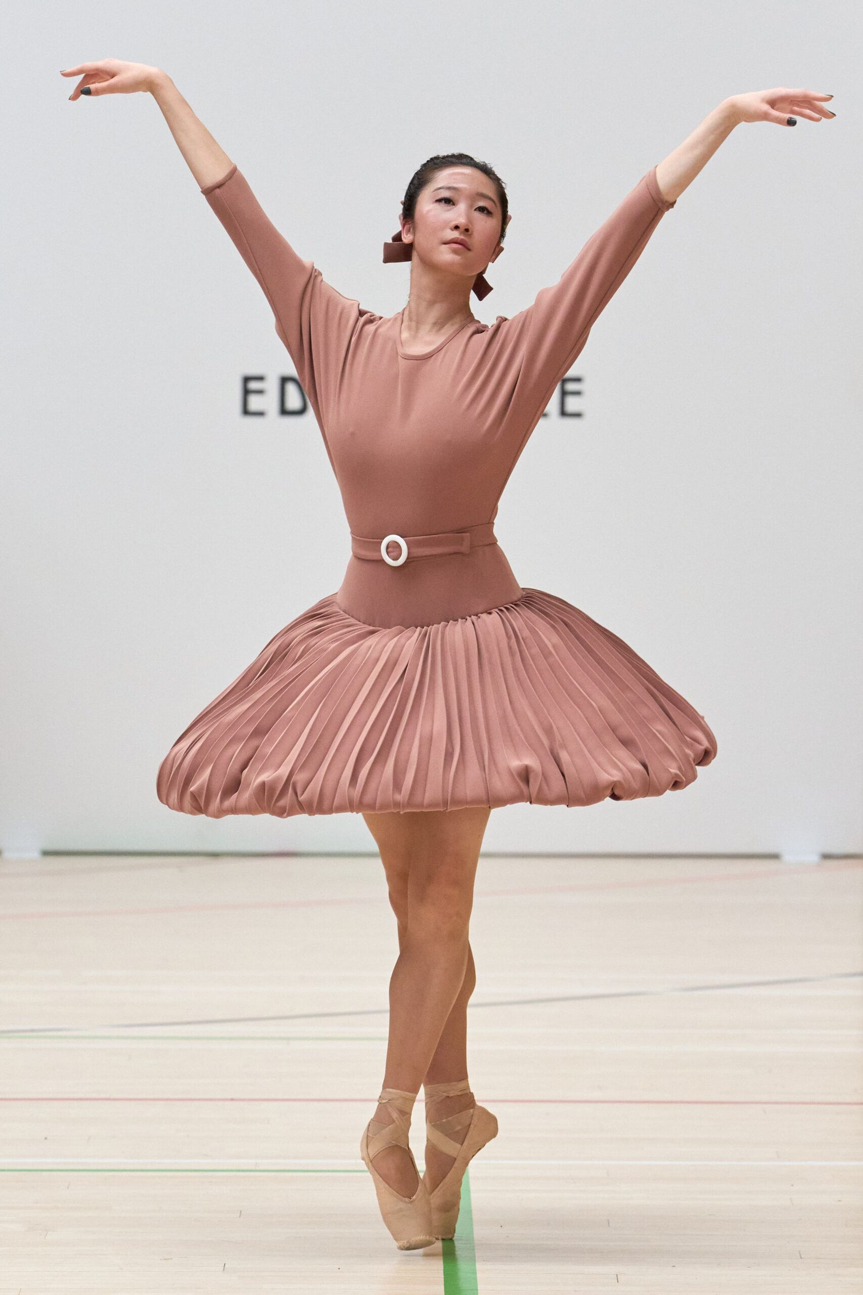 Dusty rose jersey dress with a scooped neck and long fitted sleeves. Self belt cinched with a white oval buckle at the waist. Bubble mini skirt in tight sunray pleats, puffed under at the hem. Blush satin pointe shoes with crossed ribbons. Midafternoon on a gym floor, waiting for the pianist to begin again.