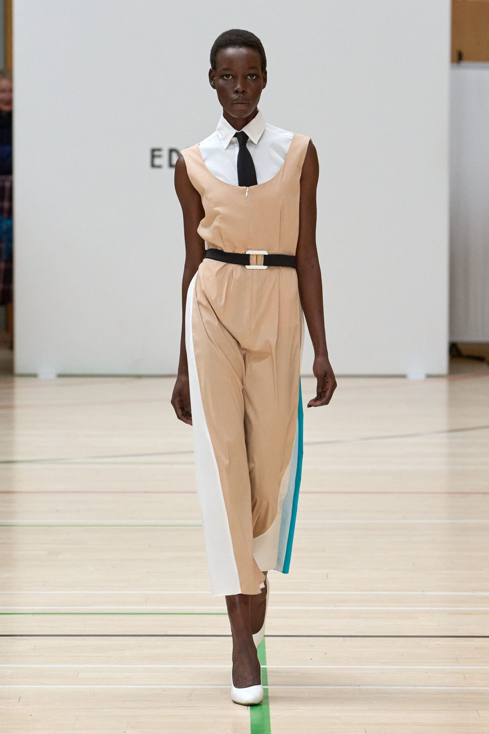 Crisp white sleeveless shirt with a pointed collar and black tie. Apricot crepe jumpsuit with a scoop neckline and white side panels. Black belt with a chunky ivory rectangular buckle. White pumps with a squared vamp. Crossing a school gym floor after hours, still dressed for the meeting.