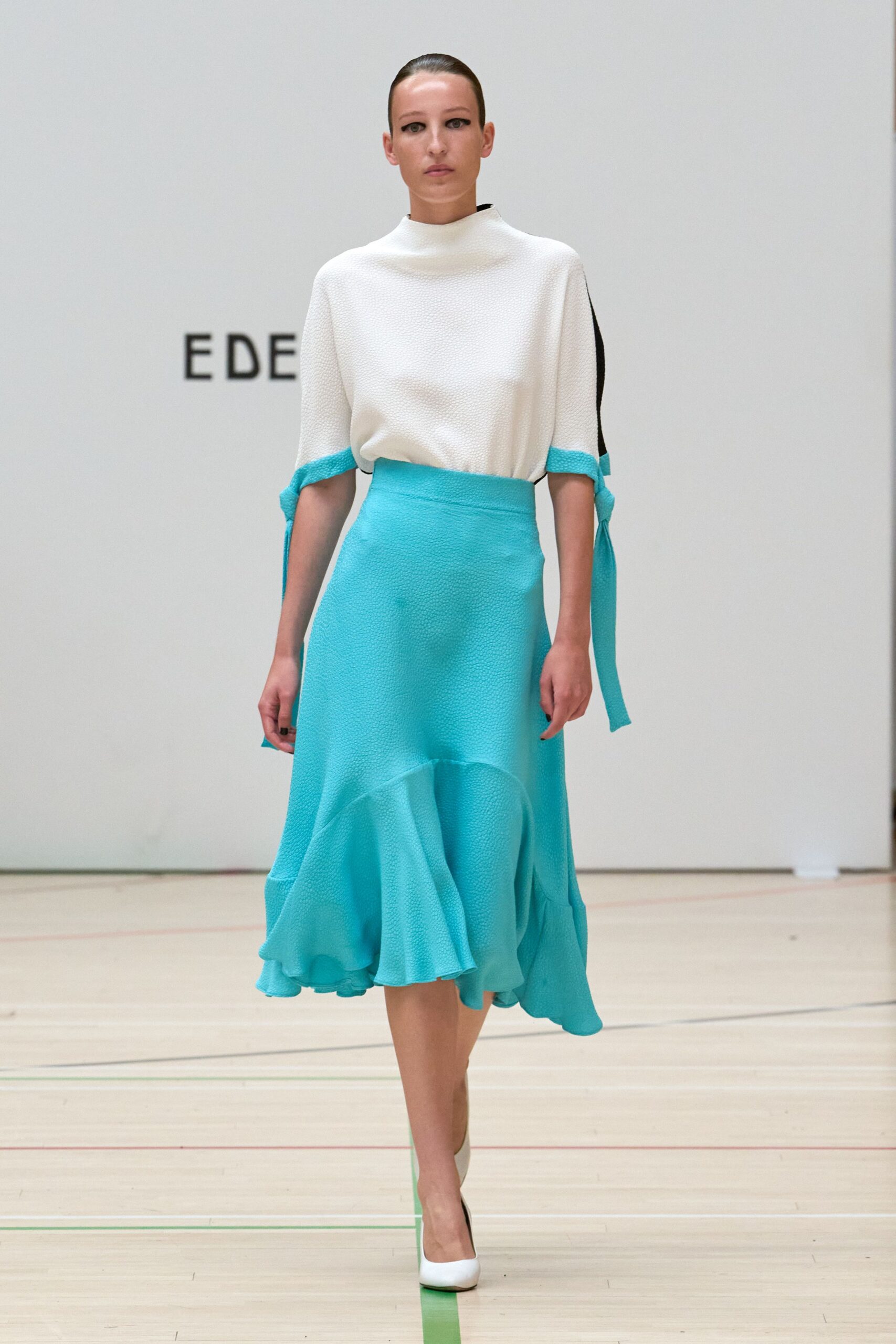 White pebble-textured top with a funnel neck and tied elbow slits. Aqua crepe skirt, high-waisted with a dropped flounce kicking forward. White pumps with a green-edged sole. Crossing a civic atrium before the fountain switches on.