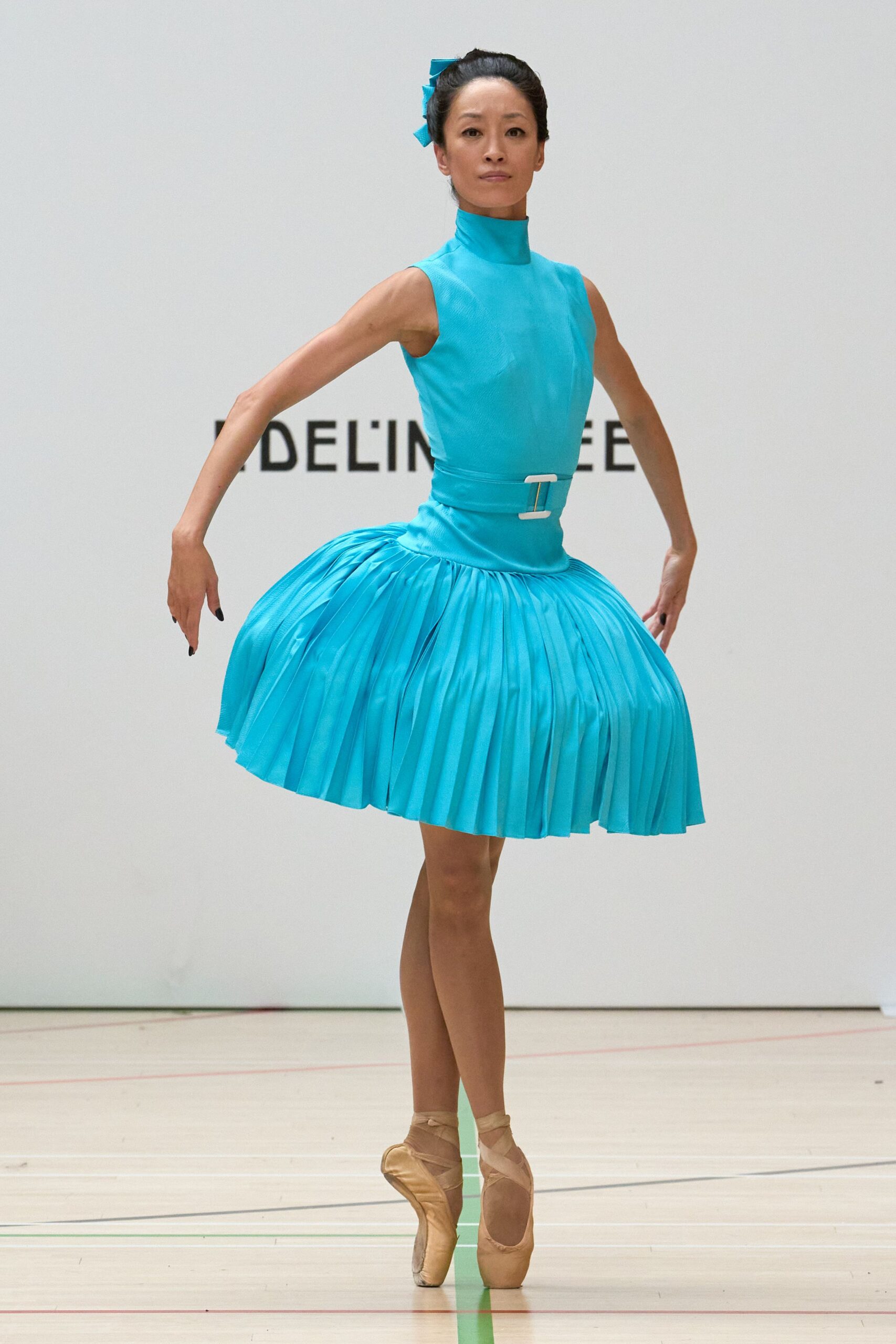 Turquoise mock-neck dress with a white rectangular belt buckle and ballooned pleated skirt. Matching turquoise ribbon bow pinned into the bun. Blush satin pointe shoes with crisscrossed ribbons at the ankle. Matinee call in a school gym, tape lines still visible on the floor.