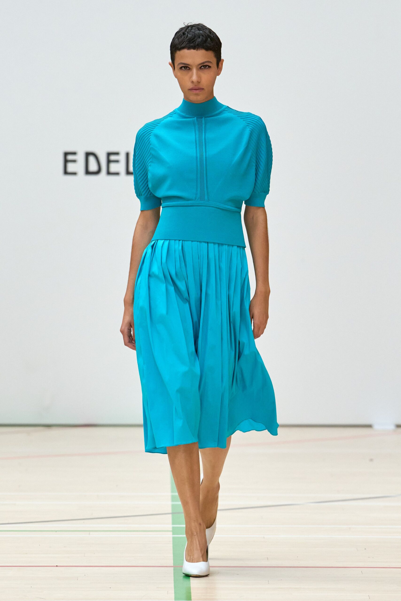 Turquoise knit top with ribbed mock neck and finely pleated sleeves. Matching skirt in fluid crepe, knife pleats falling from a wide yoke. White pumps with rounded vamps. Walking the echoing corridor outside a civic pool before it opens.
