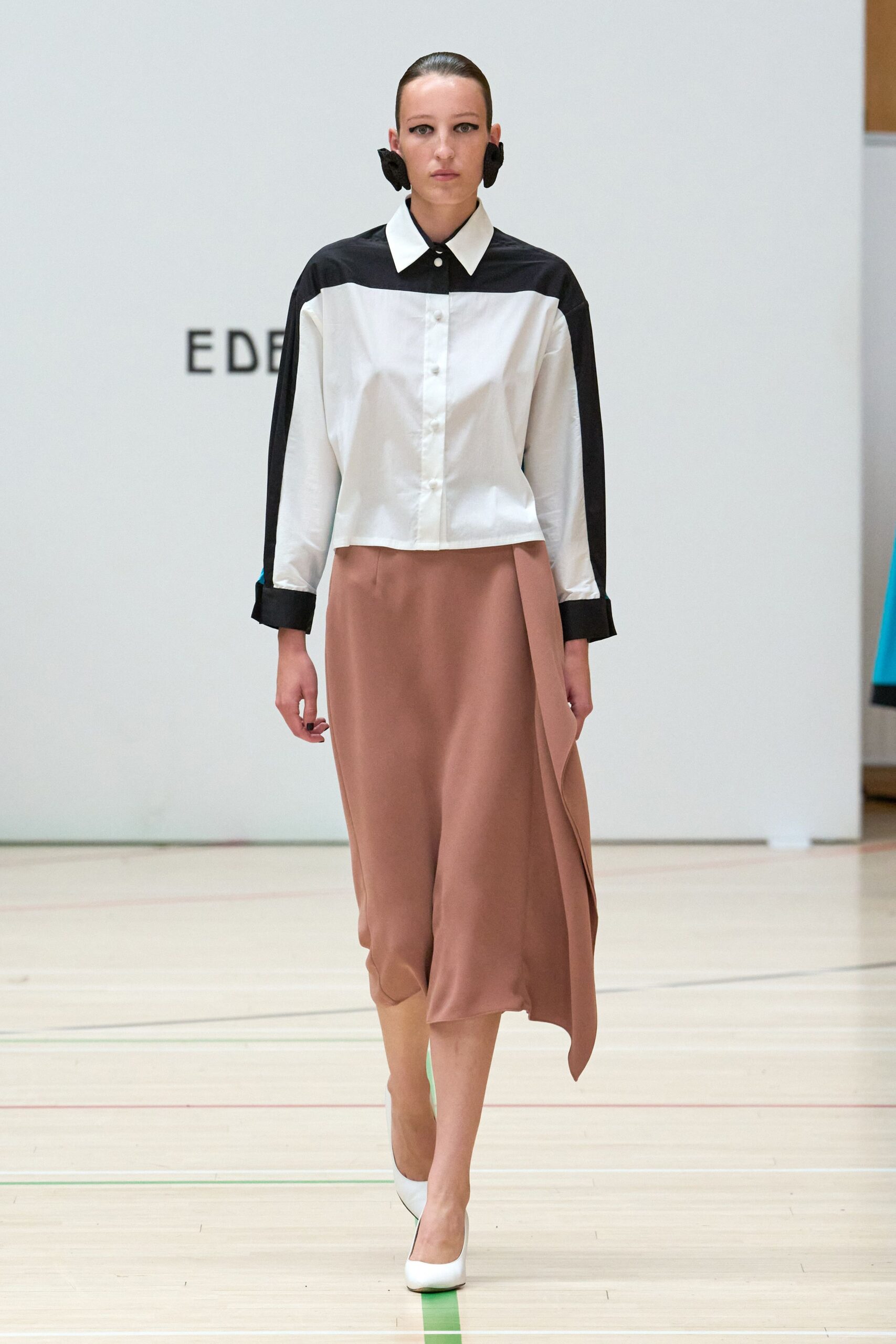 Black fabric rosettes clipped broad against the ears. Cropped white poplin shirt with black yoke, collar and turned cuffs. Dusty rose satin skirt cut on the bias, one panel falling long. White pumps with a narrow black edge at the sole. Crossing a school gym floor after everyone else has gone home.