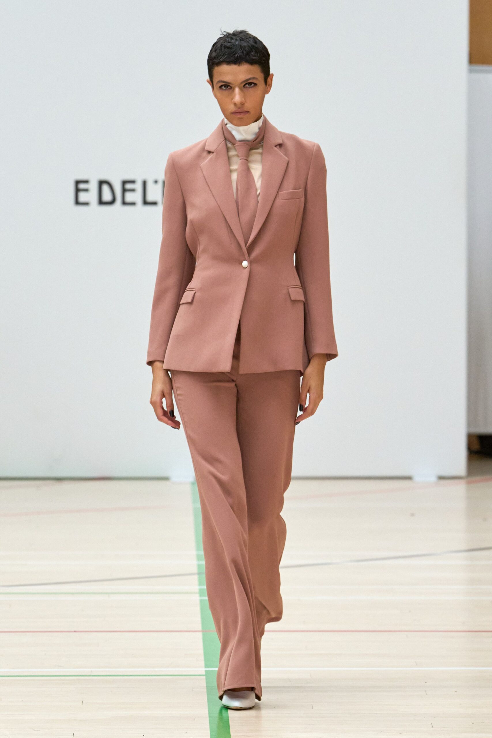 Dusty-rose tailored blazer, single pale button and flap pockets. Cream high-neck layer, sheer through the chest under the tie. Matching dusty-rose tie hanging narrow and flat. Straight dusty-rose trousers breaking softly over the shoe. White square-toe shoes. Walking into a city hall wedding before anyone has found the rings.