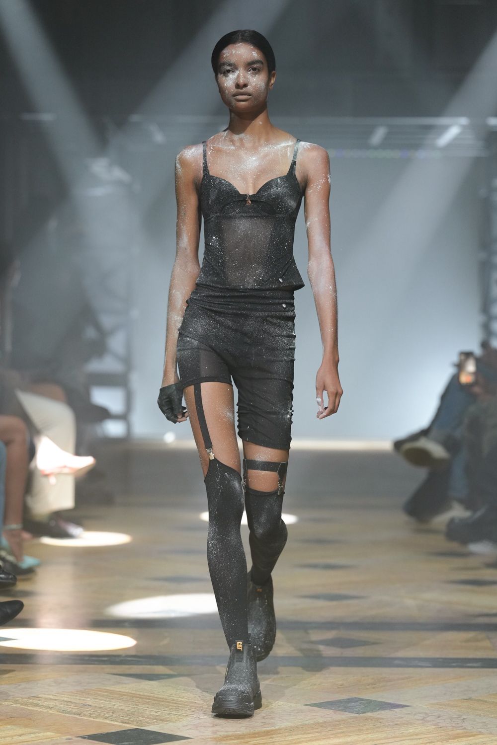 Black glitter mesh bodysuit with bra seaming and a tiny center tab. Ruched black mini pulled sideways over one hip. Single sheer thigh-high clipped with two satin garter straps. One short black glove. Glittered rubber ankle boots with a yellow pull tab. After midnight in a warehouse, talc still hanging in the lights.