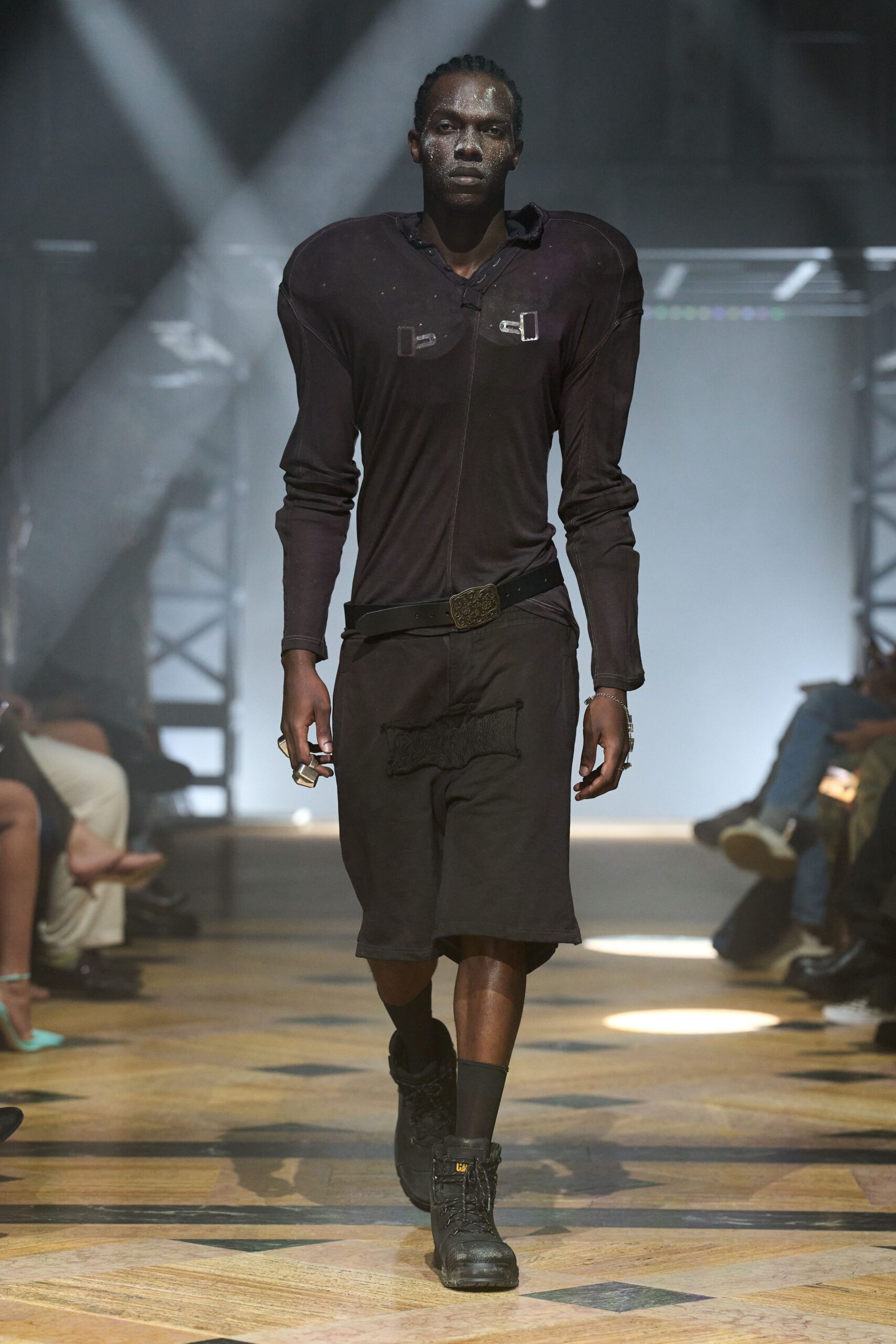 Black stretch top built with raised shoulder pads and chest buckles. Low black belt with an engraved square plaque buckle. Long black shorts, one thigh patched with furry black pile. Black mid-calf socks. Black work boots with a yellow tongue tab. Walking across a gym floor after the generator kicked back on.