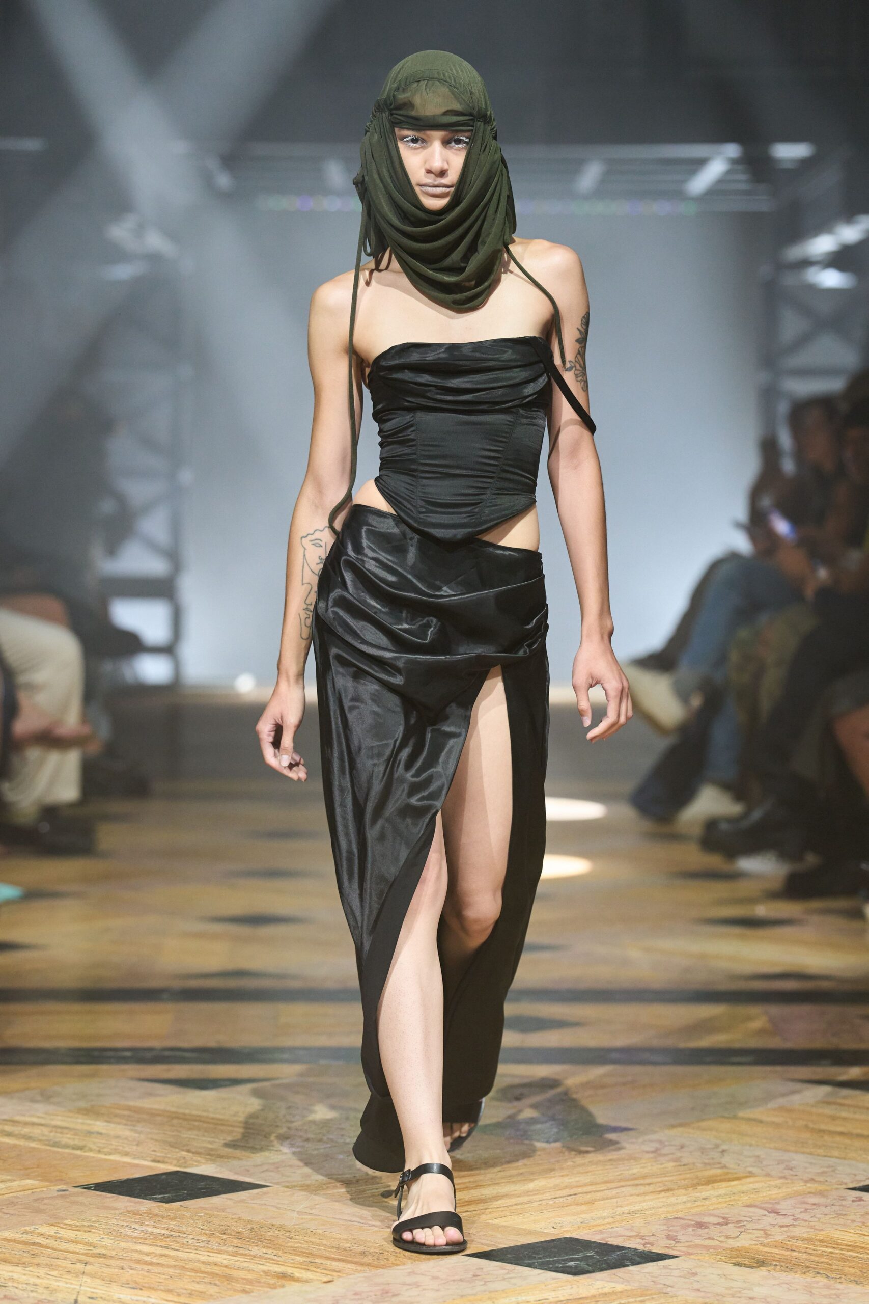 Olive jersey hood wrapped into a cowl, cords hanging past the chest. Black satin corset top, ruched bandeau edge and a sharp pointed front. Bias-cut black satin skirt slung low, one thigh exposed by a deep split. Black ankle-strap sandals. Crossing a tiled bathhouse corridor before the steam room door swings shut.