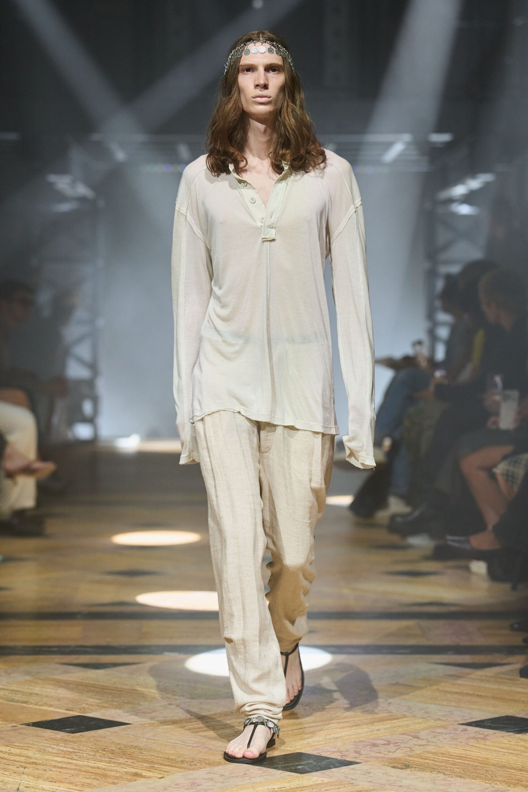 Silver coin chain draped low across the forehead. Ivory jersey henley, raw placket hanging below the third button. Loose ecru gauze trousers with a faint crinkled vertical weave. Black thong sandals with jangling silver charms at the ankle. Walking back from the beach through a hotel lobby still cool from dawn.