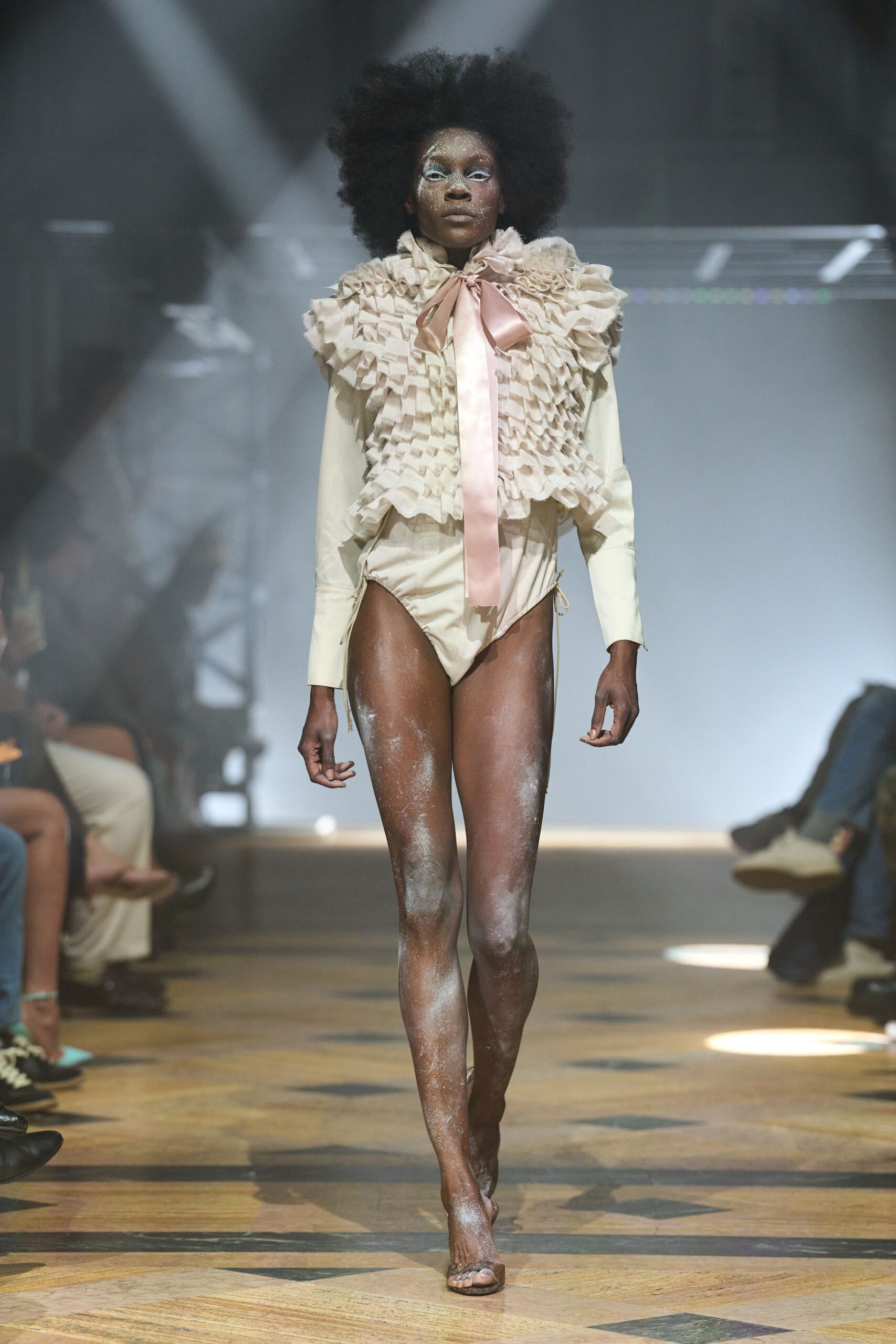 Cream organza bib built from tight box pleats and frothing ruffles. Blush satin bow hanging long past the sternum. Ivory long-sleeve bodysuit with a high-cut leg and side ties. Clear heeled mules, toes pushed to the edge. Powder room haze after the costume trunk has been thrown open.