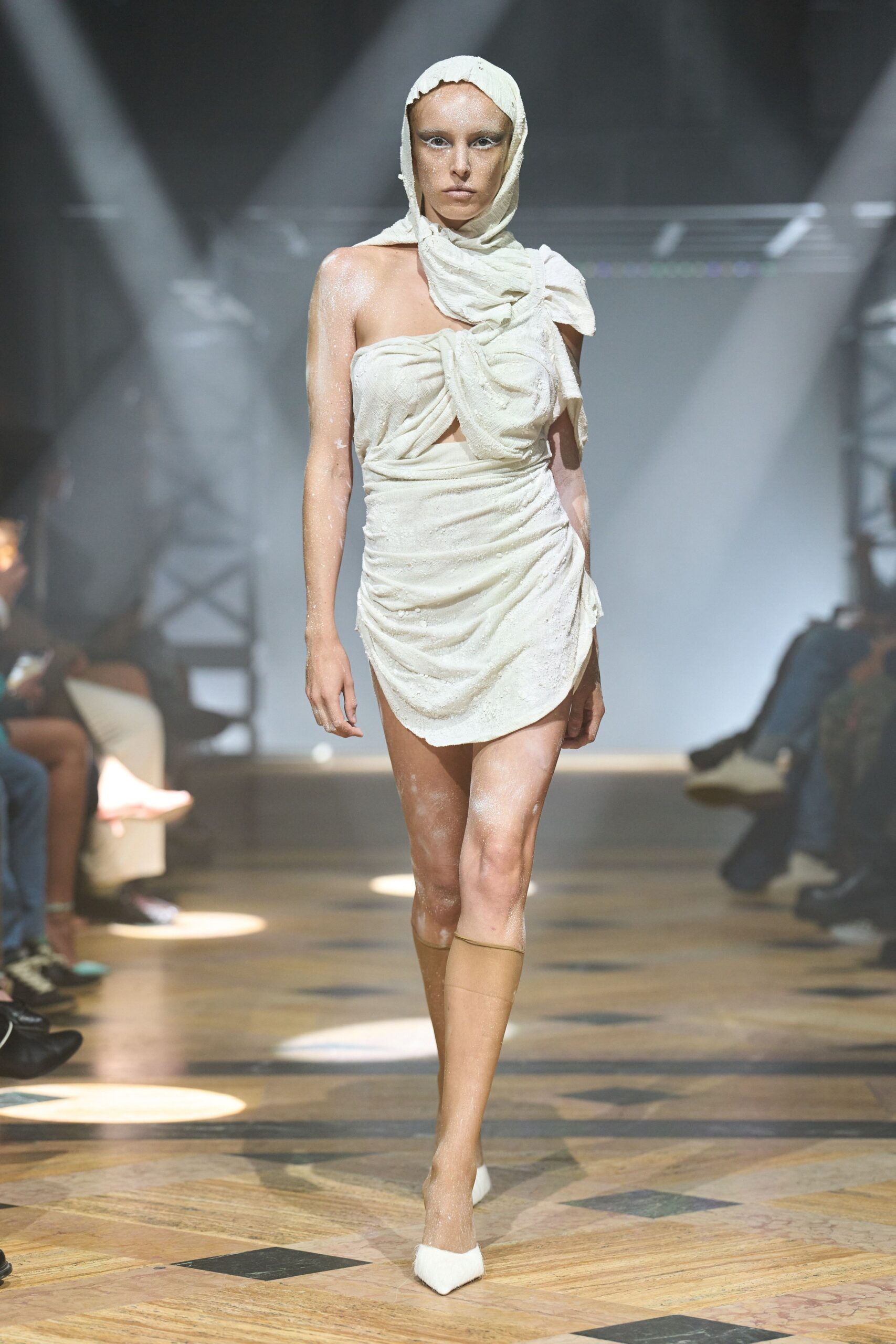 Gauzy ivory hood with a ragged edge framing the forehead. Ivory jersey mini, twisted into a bowlike bandeau and ruched hip drape. Sheer nude knee-highs, one sagging slightly below the knee. White pointed pumps tipped with a fuzzy mohair cap. Morning after the spa, robe traded for whatever was still warm.