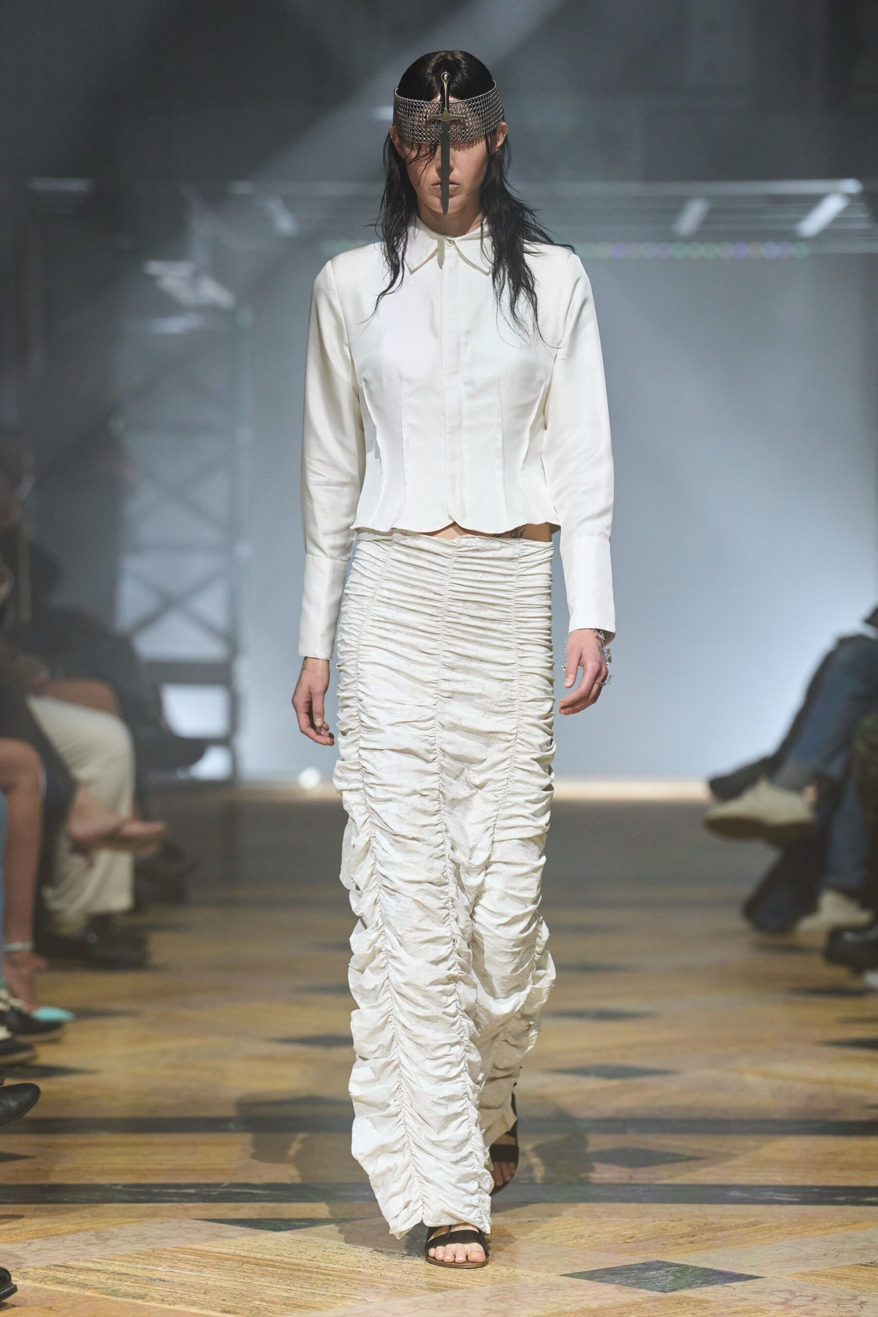 Silver chainmail visor pinned with a long dagger down the face. Ivory fitted shirt with a concealed placket and pointed hem. Crushed satin column skirt densely ruched into vertical gathers. Black thong sandals. Church hall air after rain, walking in with one earring missing.