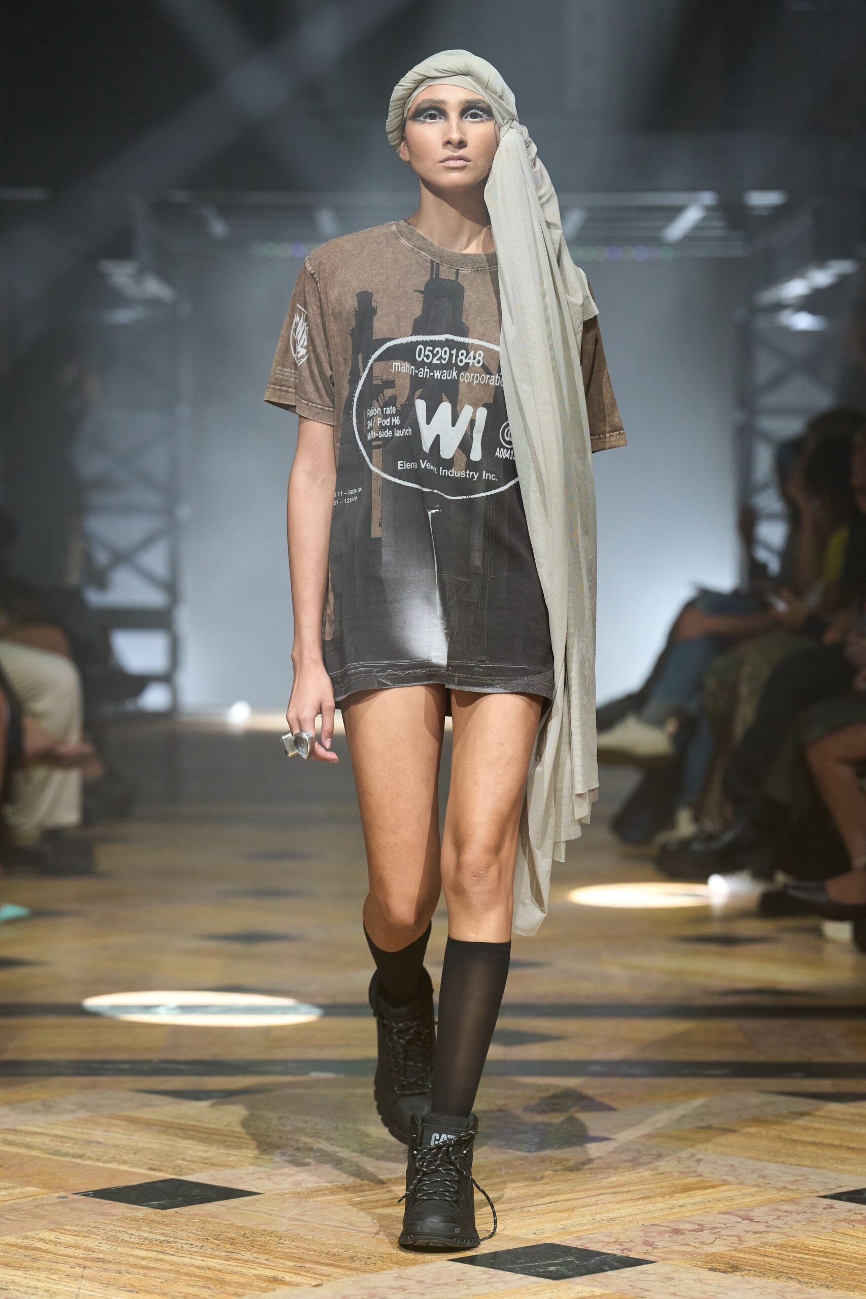 Pale gauze turban wrap with one long panel falling past the hip. Oversized washed brown graphic tee, white stencil print and raw-looking hem. Chunky clear resin ring. Sheer black knee socks. Black lace-up combat boots, one loose lace tip dragging. Crossing a train platform at dawn with last night's makeup still on.