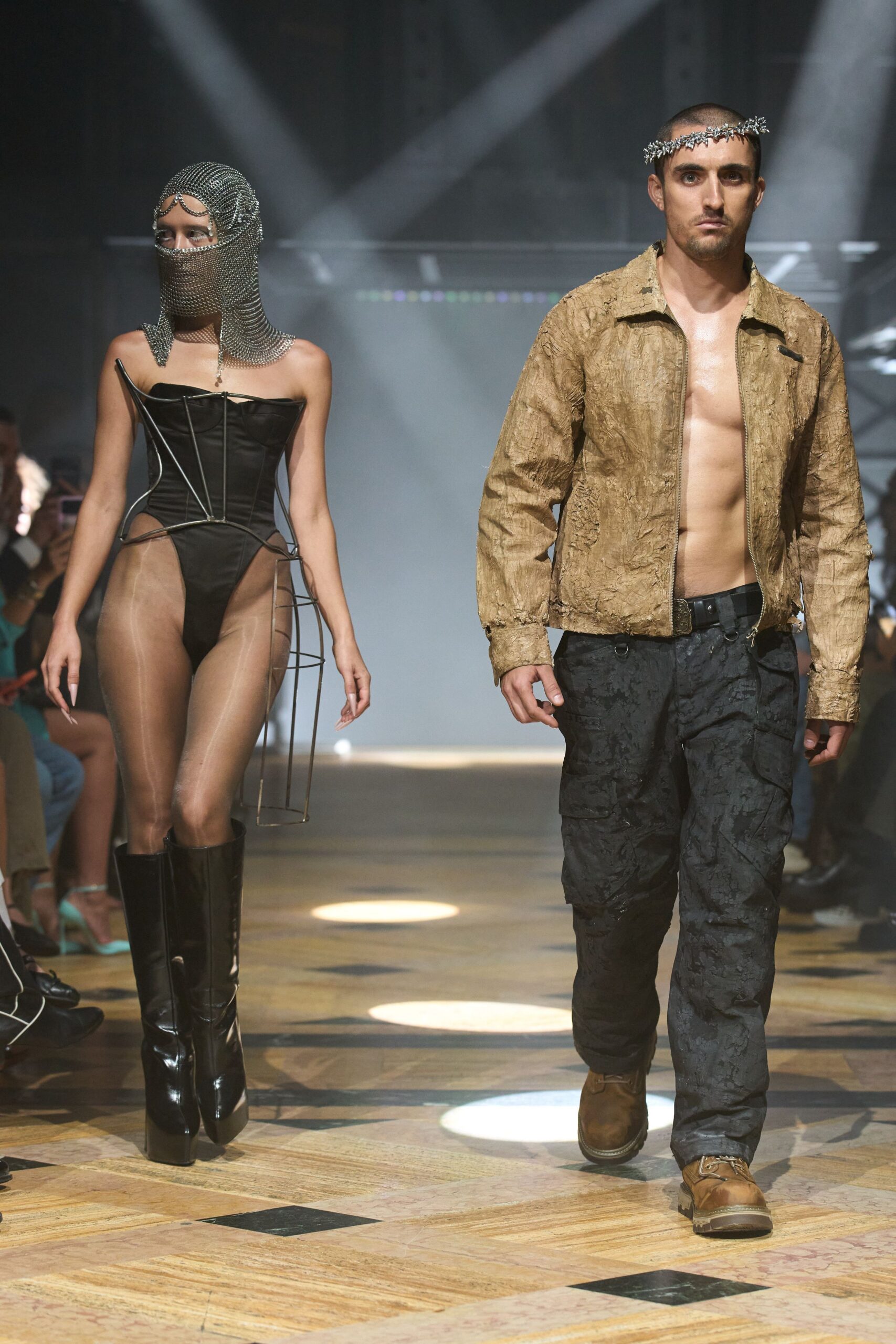 Silver thorn circlet worn low across the buzzed forehead. Raw khaki jacket, papery and crumpled, zipped open to the sternum. Black cargos in blistered jacquard, D-rings hanging off the waistband. Mud-brown work boots with thick lug soles. After the bonfire wedding, still crossing the field for the cars.