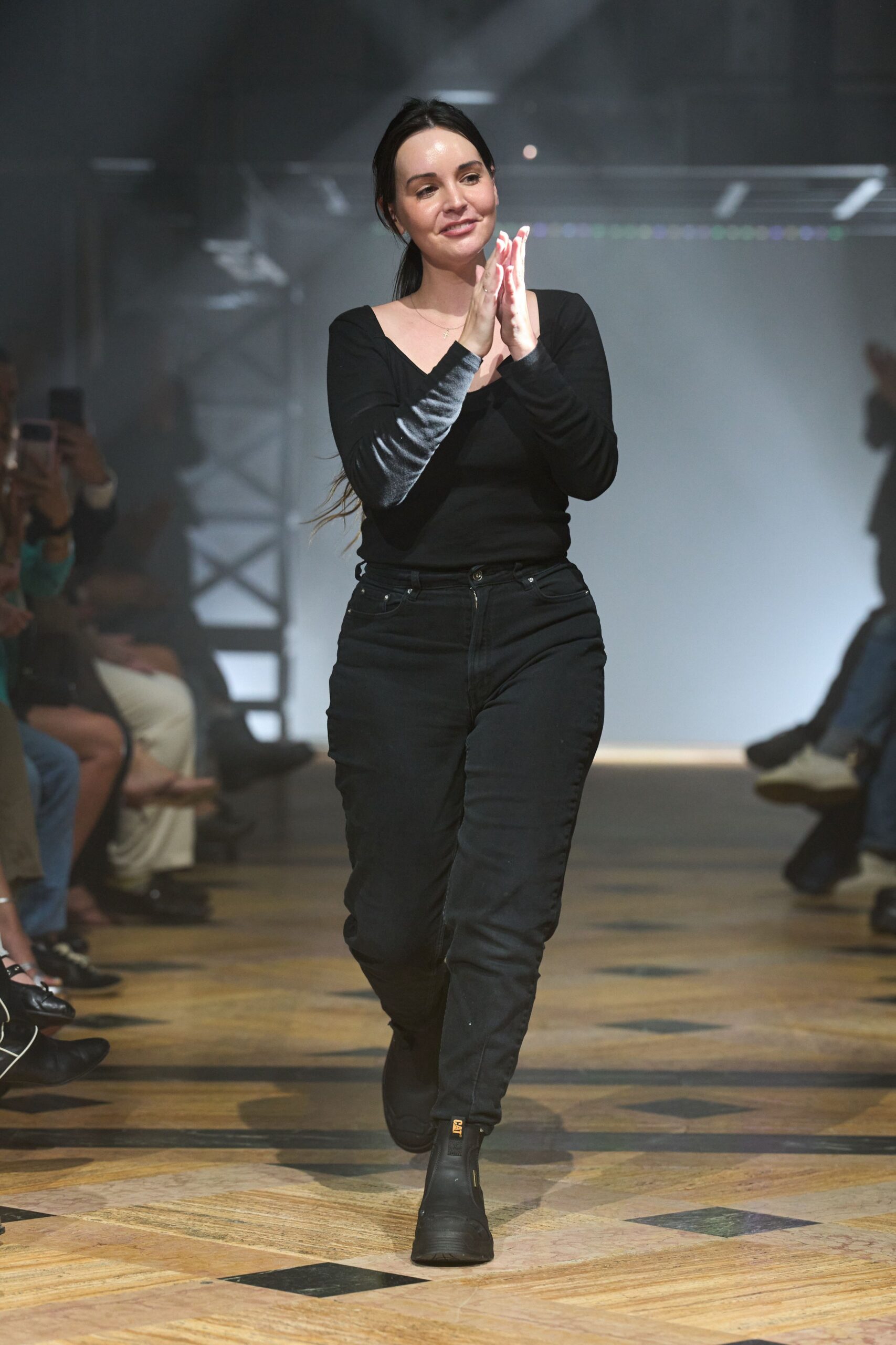 Black jersey scoop-neck top, sleeves pushed and shining at the forearms. High-rise black jeans with riveted coin pocket and a centered zip fly. Matte black work boots with a yellow logo tab at the shaft. Clapping at the end of a warehouse fundraiser, floor still dusty from setup.