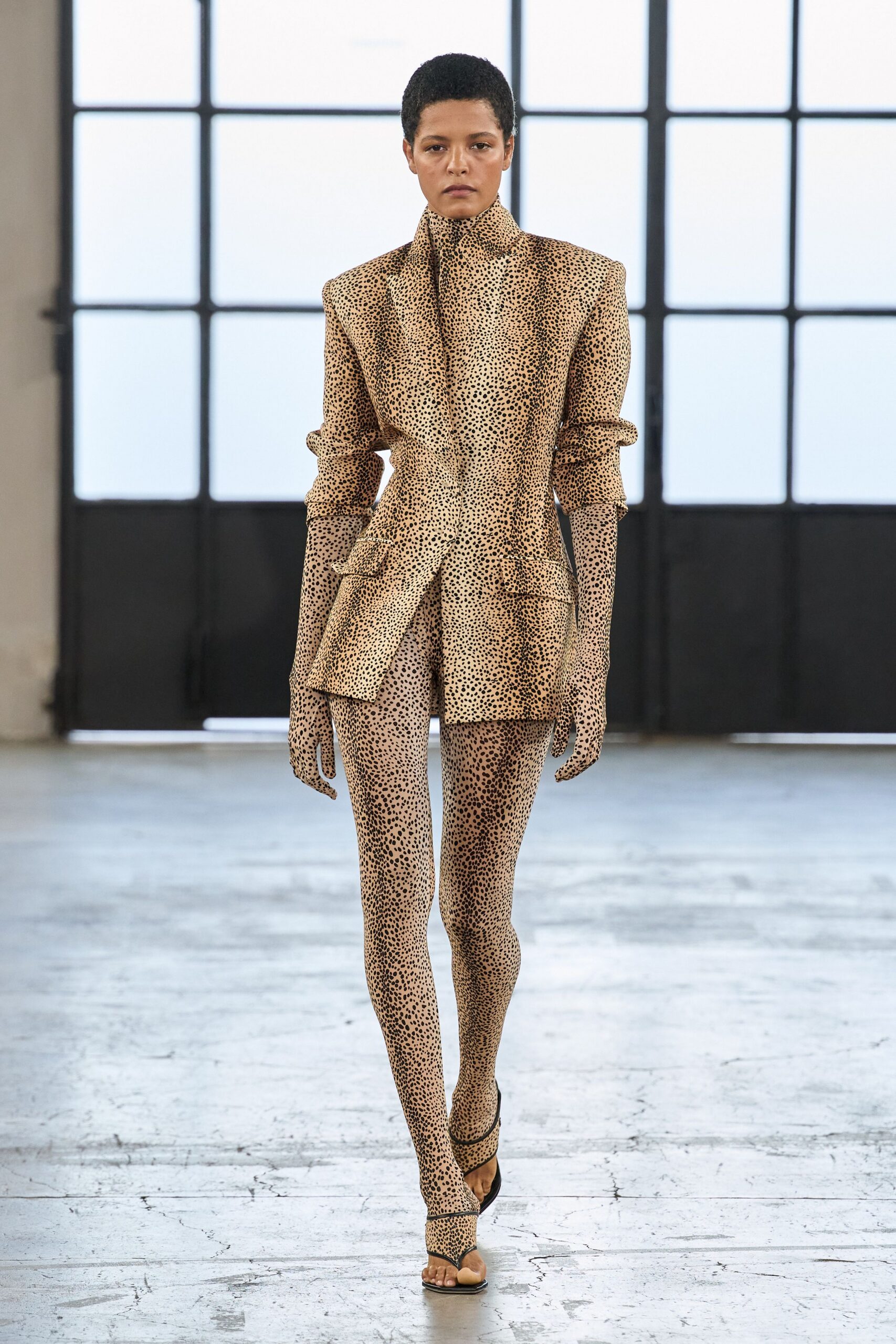 Sand suiting jacket speckled like cheetah, collar rising to the jaw. Matching opera gloves, one finger hanging slightly longer. Second-skin leggings in the same dense spotted stretch. Black toe-ring sandals with a narrow curved strap. Crossing an empty loft before the estate agent unlocks the next room.