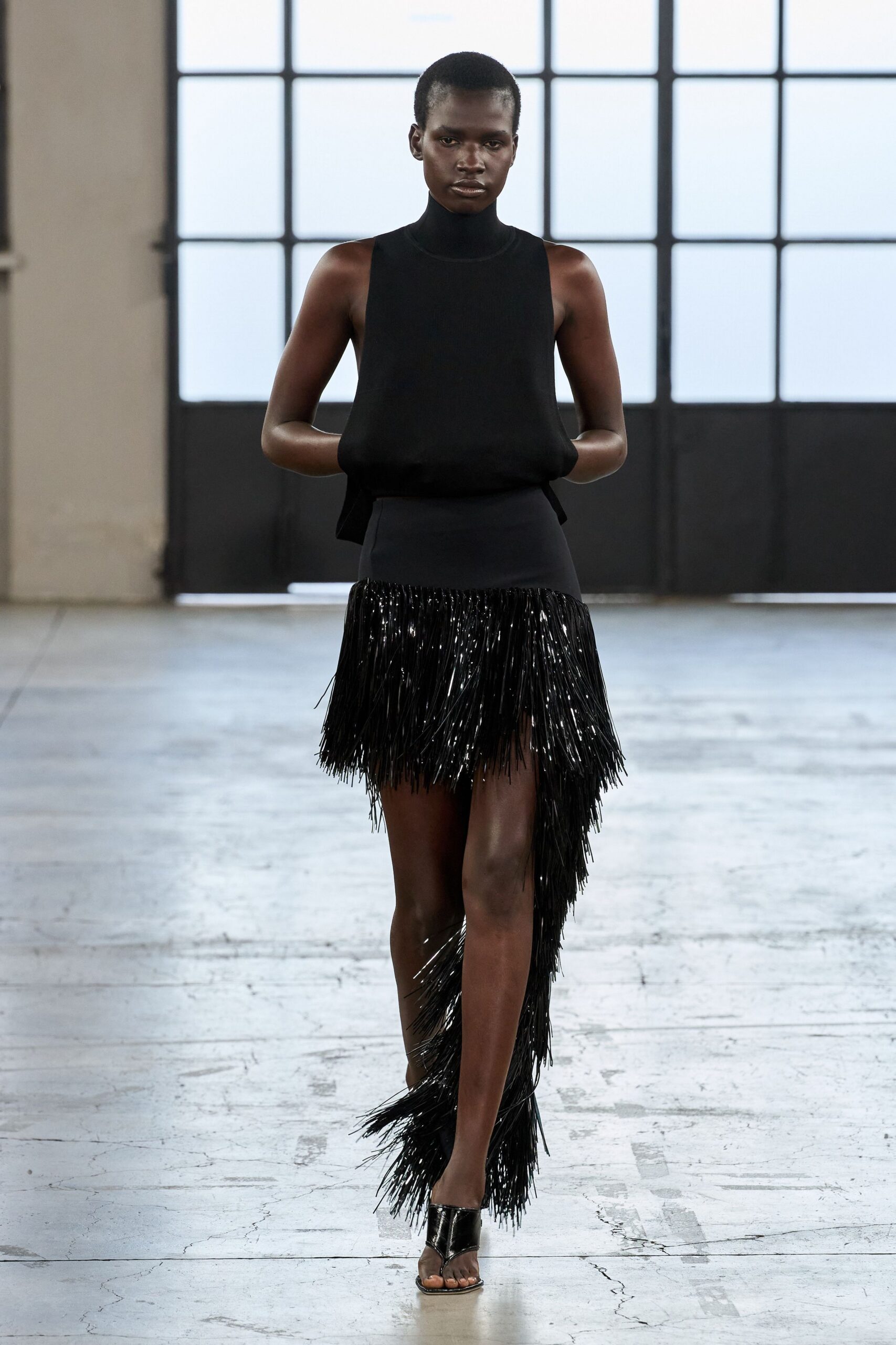 Black knit shell with a snug funnel neck and dropped armholes. Asymmetric black skirt, lacquered fringe swishing from a plain hip yoke. Patent thong mules with a sharp V over the foot. Walking into a hotel atrium while the piano tuner is still working.