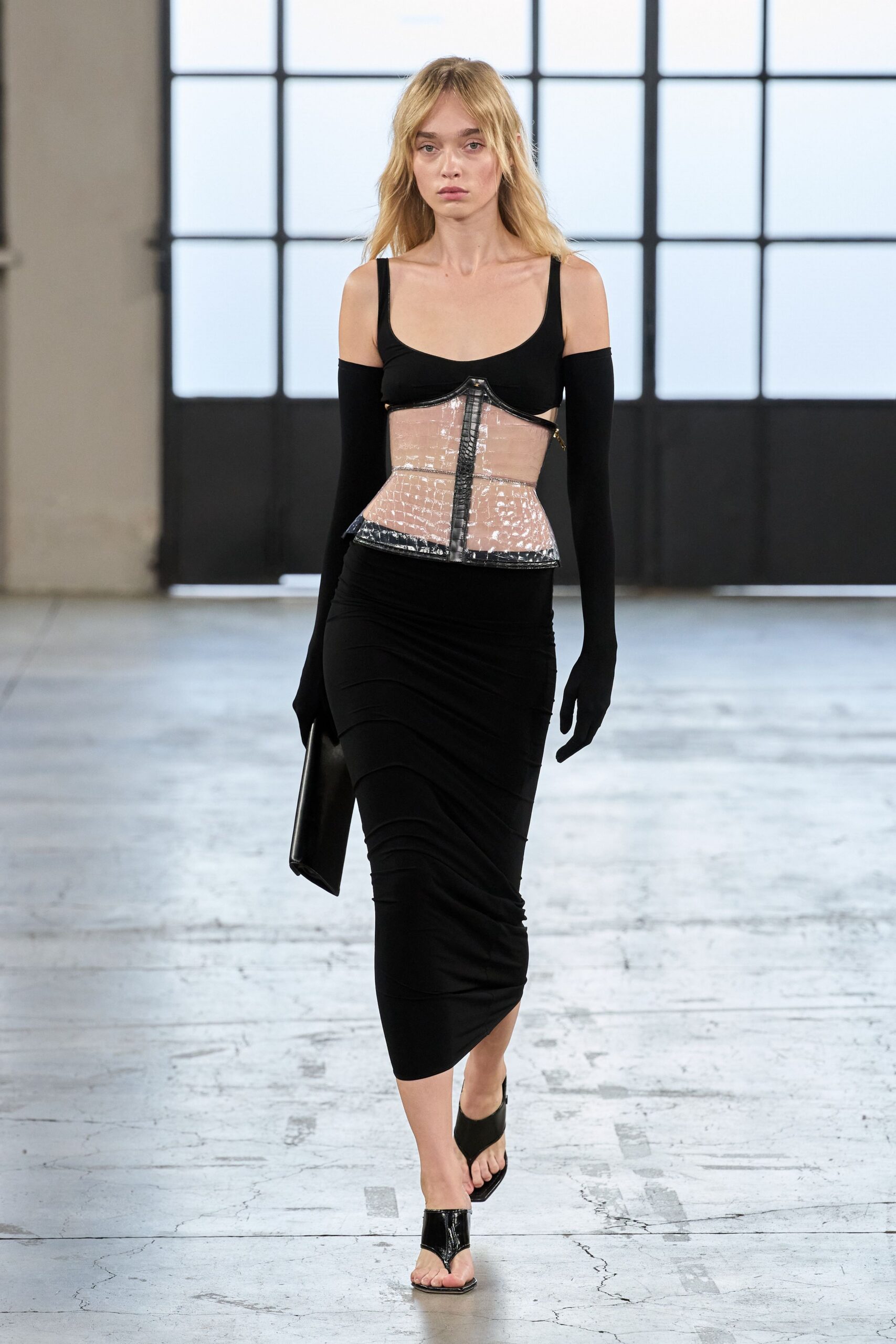 Black scoop-neck top with detached opera sleeves. Clear vinyl corset, black seamed and peplumed at the waist. Black jersey midi skirt, ruched into diagonal ripples. Flat black pouch tucked under one arm. Black patent thong mules. Crossing a near-empty terminal after checking the bag twice.