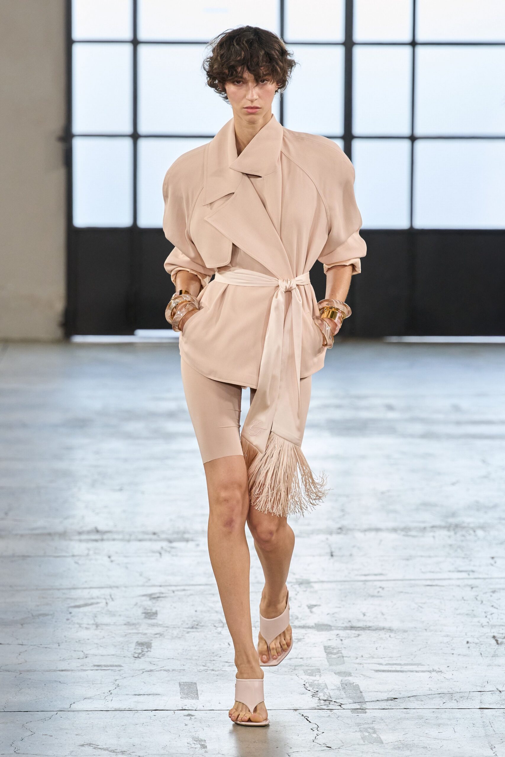 Blush satin wrap jacket with broad lapels and rolled sleeves. Self-tie sash knotted low, one end finished in long fringe. Second-skin nude cycling shorts cut to mid-thigh. Stacked translucent cuffs with one flash of gold metal. Powder pink thong mules. For crossing a silent hotel lobby just after the pool closes.