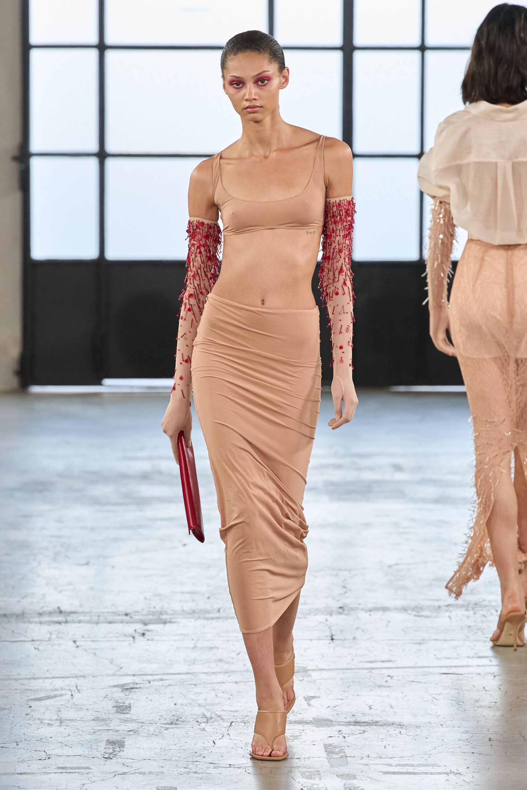 Nude stretch bralette with a low scoop and narrow straps. Sheer nude mesh sleeves drenched in red beaded fringe. Nude jersey midi skirt, ruched into diagonal drag lines. Slim oxblood clutch tucked flat in one hand. Barely there nude sandals with a wrapped ankle strap. For crossing a hotel lobby still wearing last night's makeup.