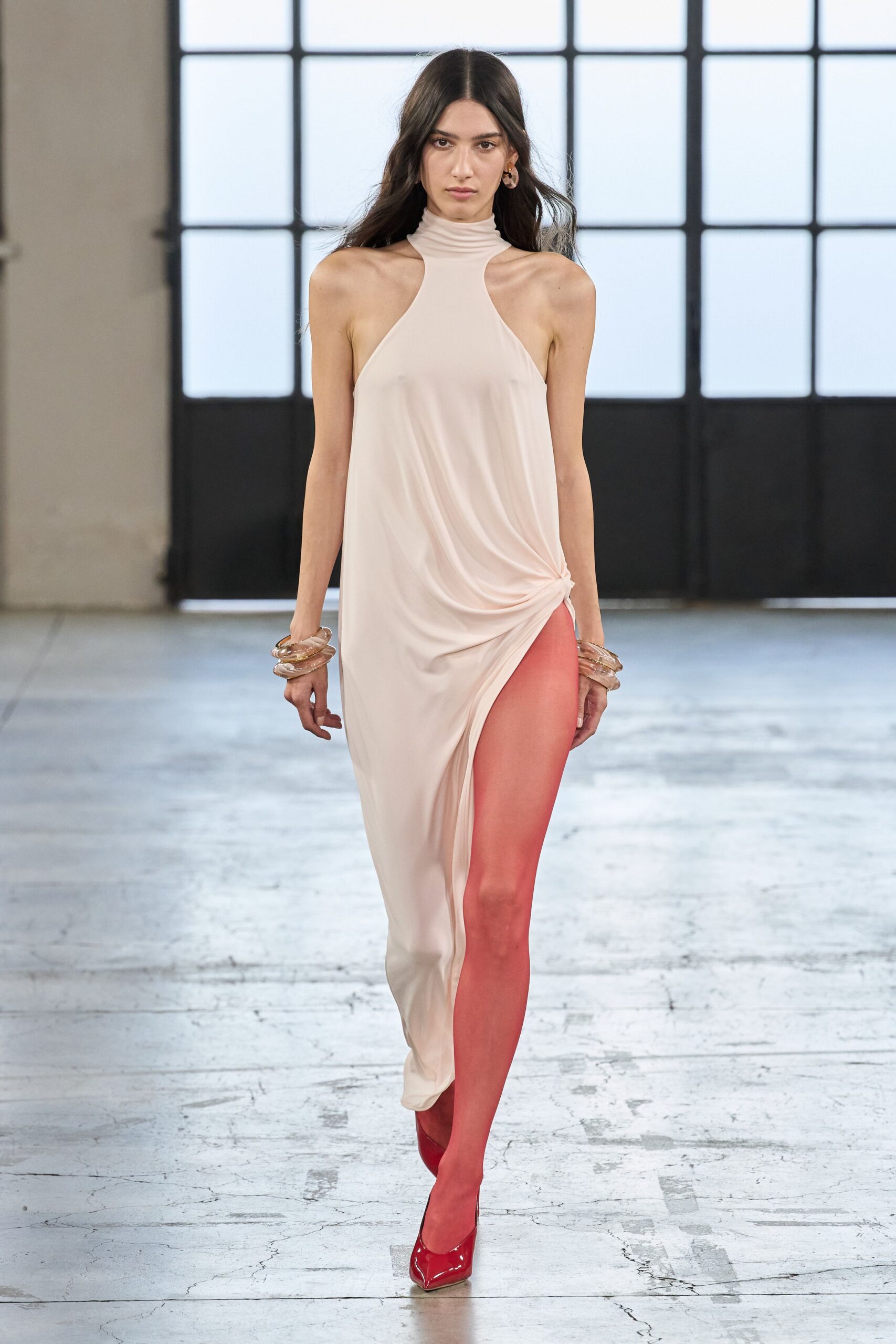 Blush jersey halter dress with a rolled neck and side knot. Tomato-red tights exposed by the thigh-high draped split. Pointed red patent pumps. Stacked translucent resin bangles at both wrists. Small amber hoop earrings. Crossing a sunlit atrium after changing in too much haste.